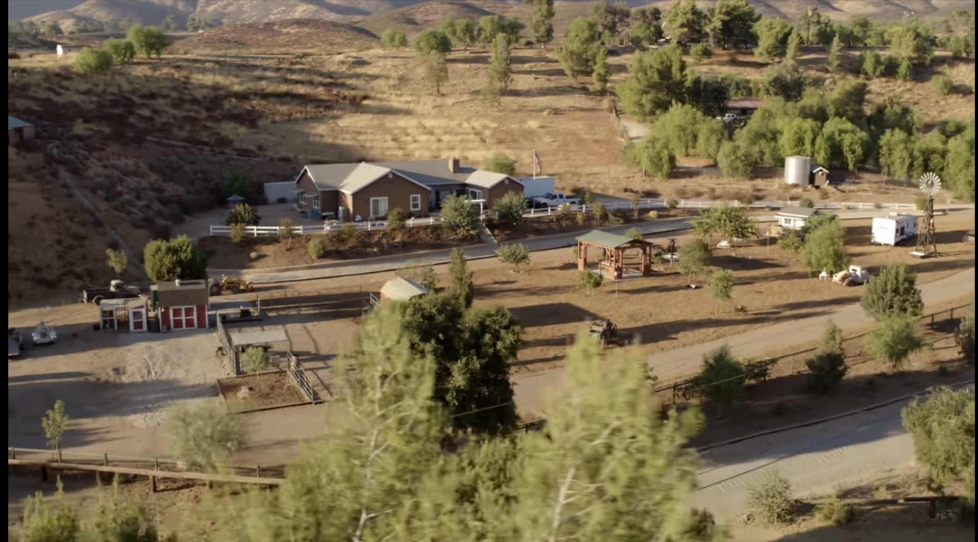 The image shows a rural property consisting of a house, several outbuildings, and fenced areas, surrounded by hills and greenery.