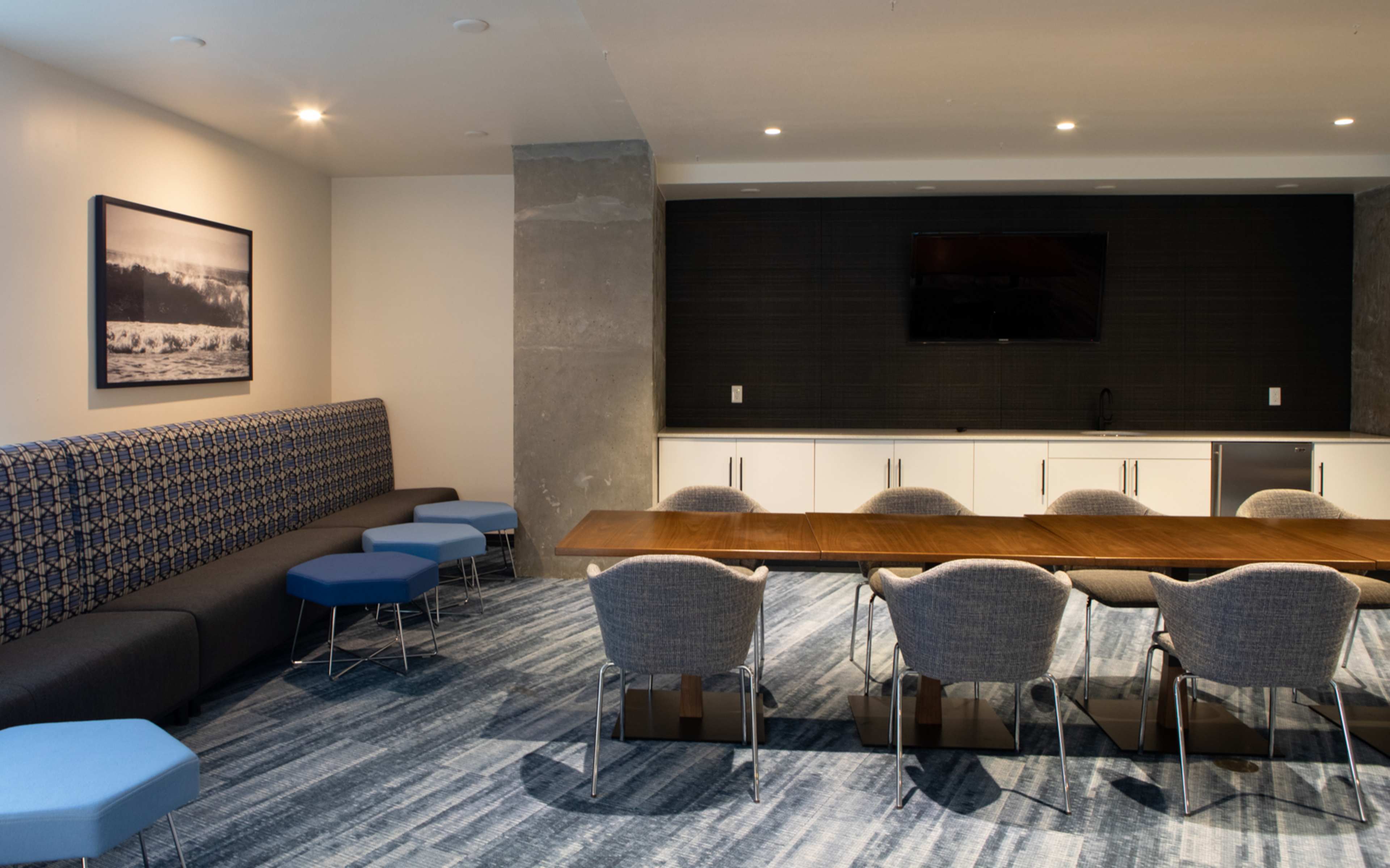 A modern meeting room featuring a long wooden table with several chairs, a blue upholstered bench, and a large wall-mounted television.