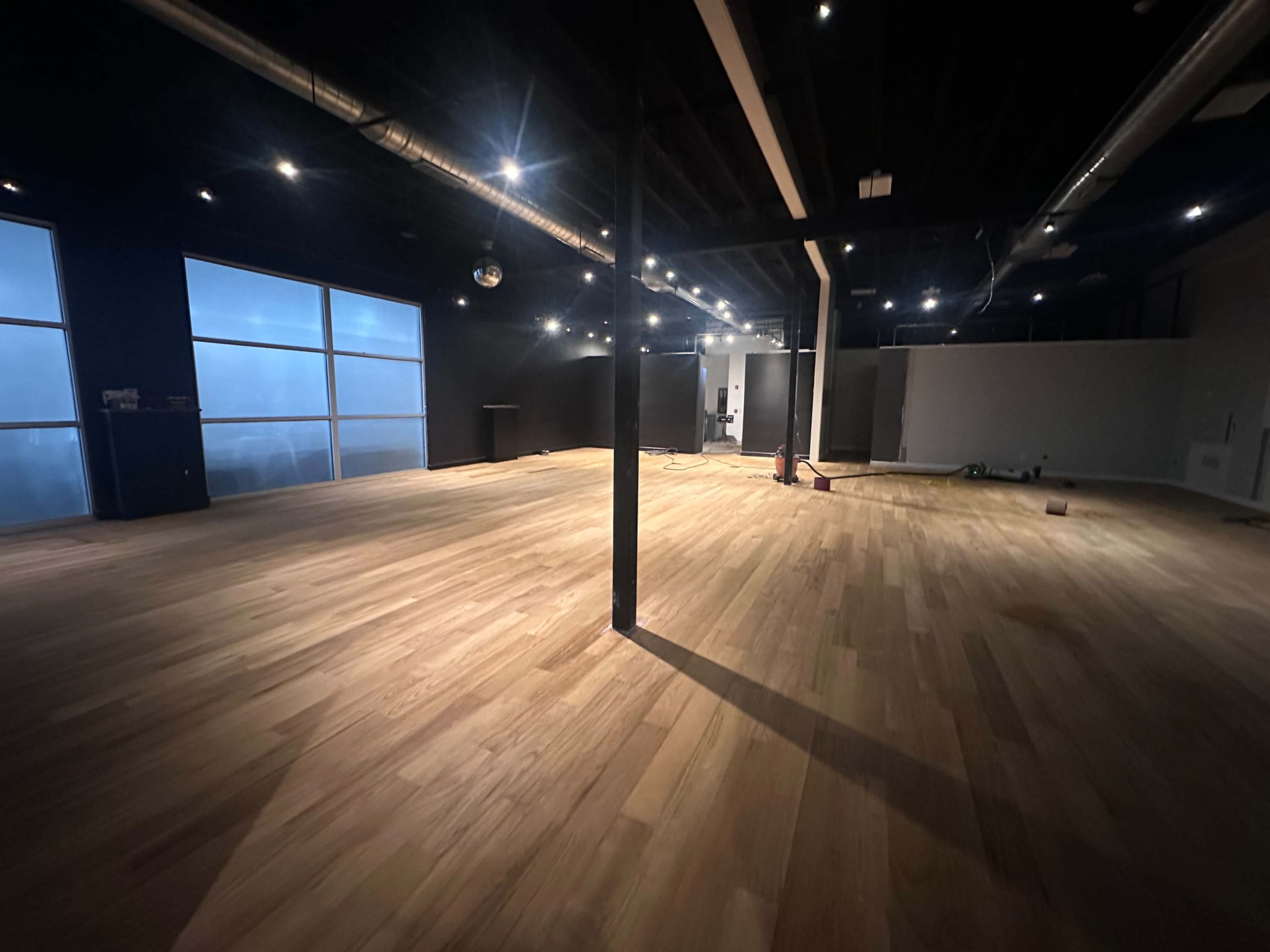 The image shows an unfinished interior space with wooden flooring, black walls, and large windows allowing natural light to enter.