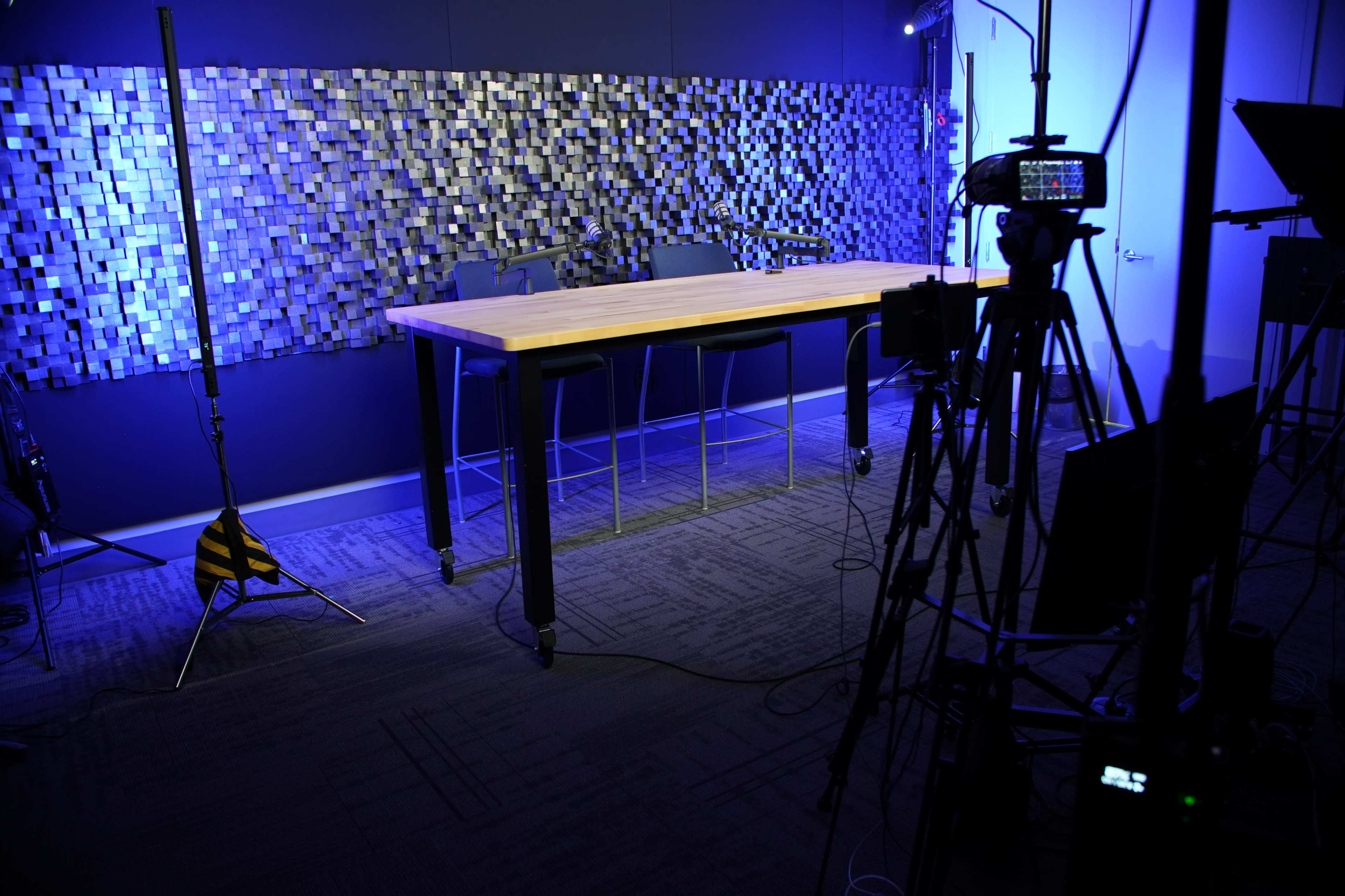 A brightly lit studio setup features a wooden table and chairs against a textured backdrop, with various cameras and lighting equipment arranged around the scene.