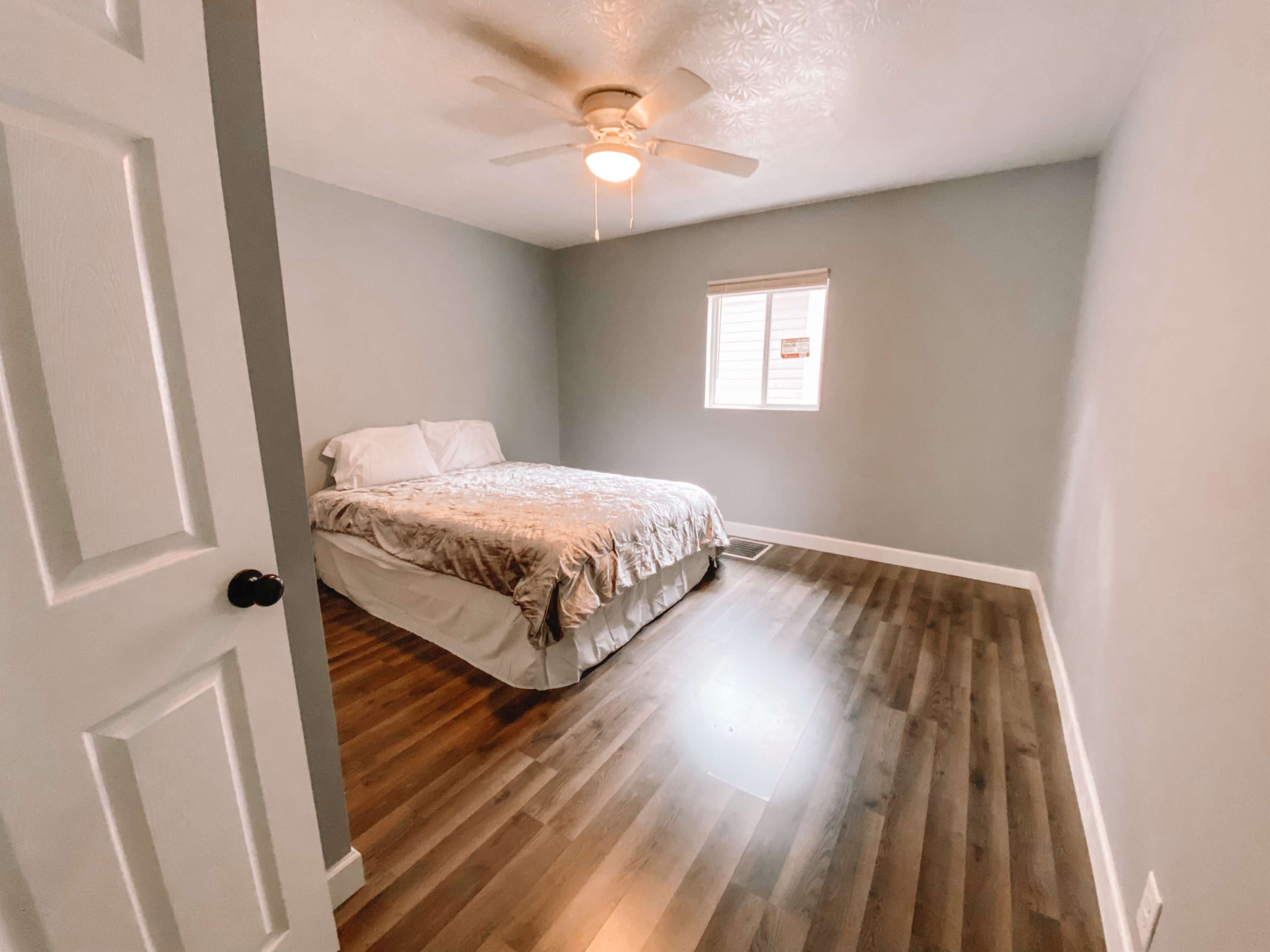 A simple bedroom features a bed with a light-colored cover, a ceiling fan, and a window with closed blinds.