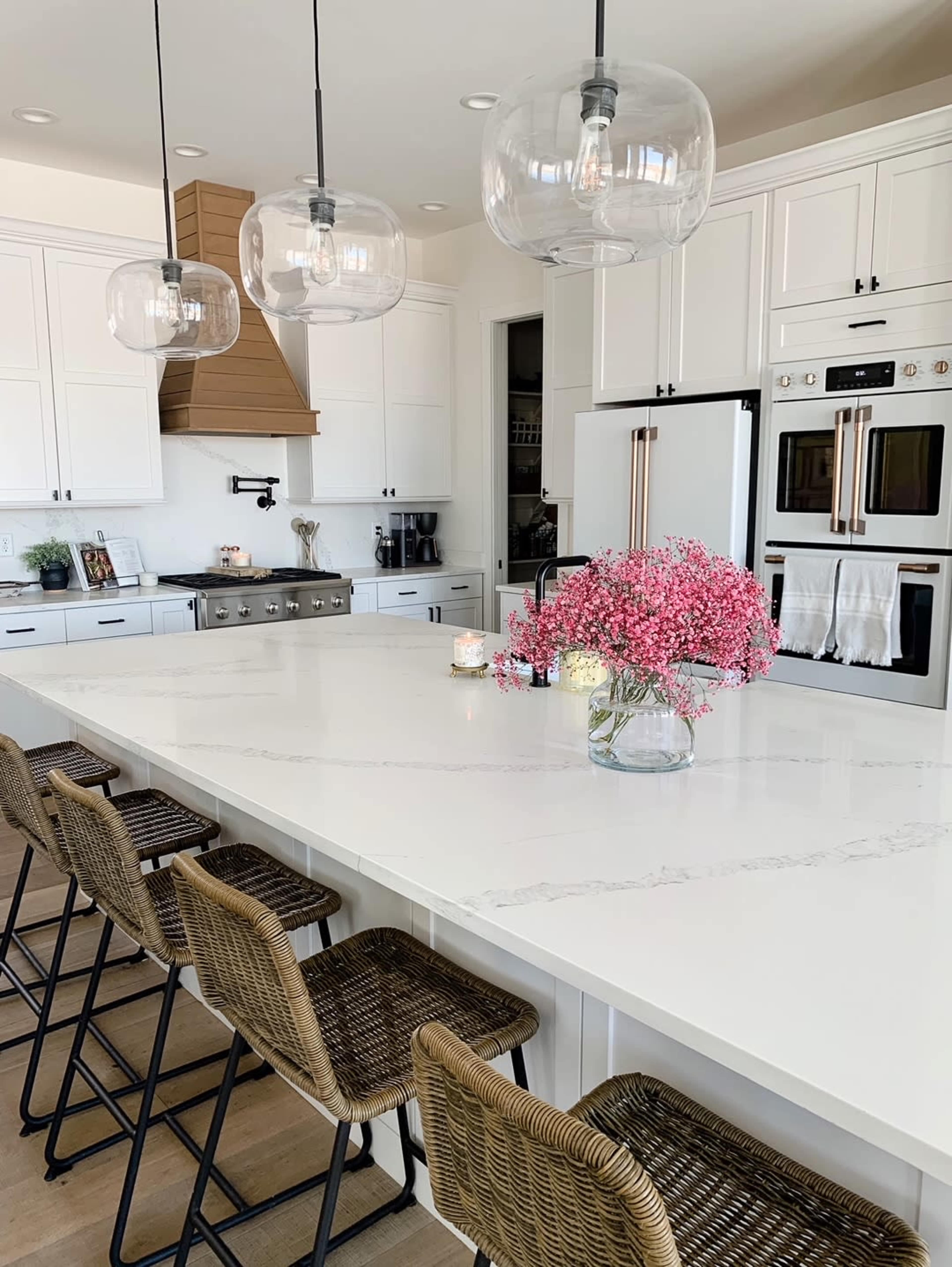 A modern kitchen features a large island with a white countertop, surrounded by wicker bar stools and illuminated by glass pendant lights.