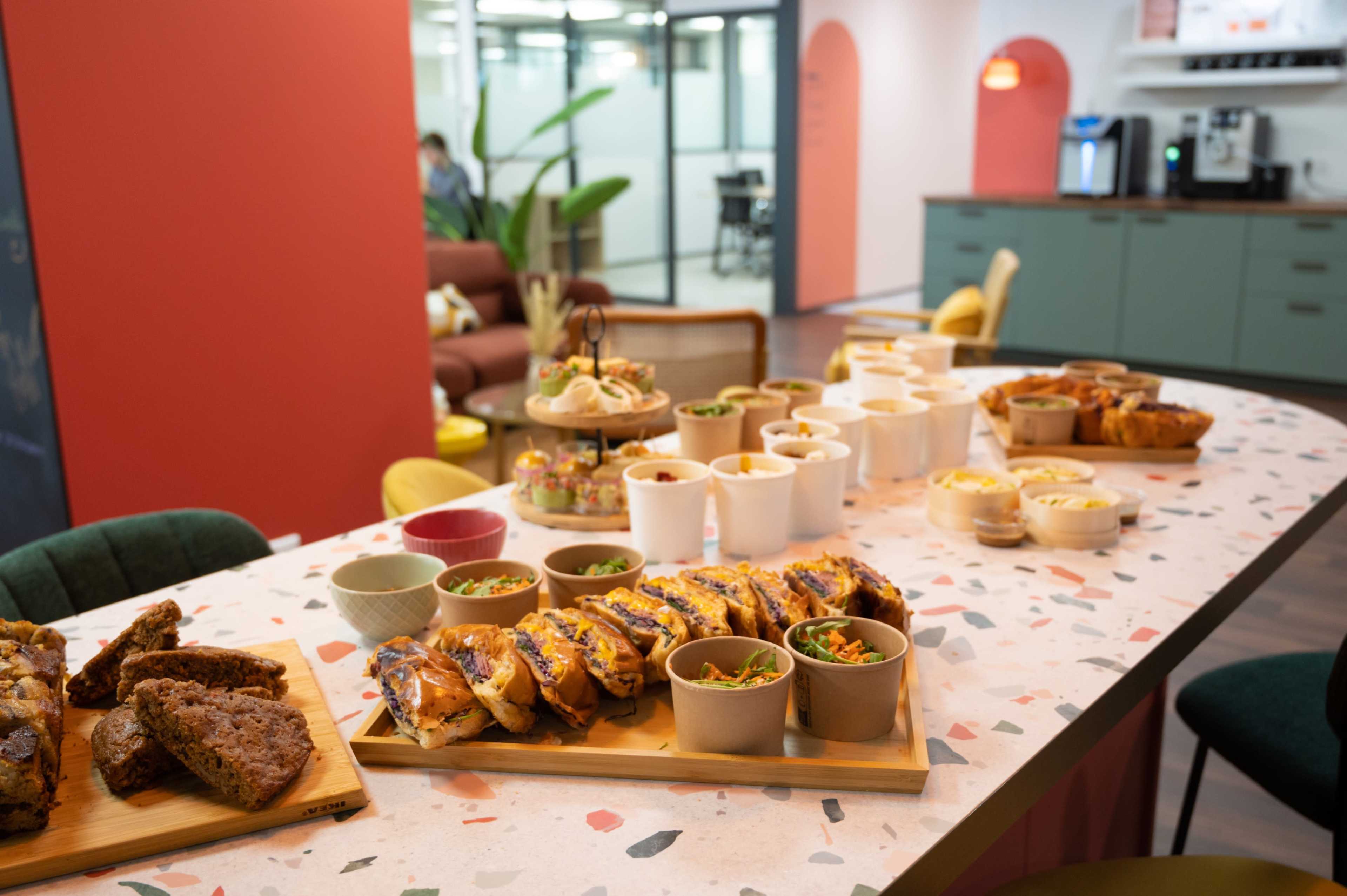 A long table is set with various food items, including breads, pastries, and small bowls of salads, in a bright office space.