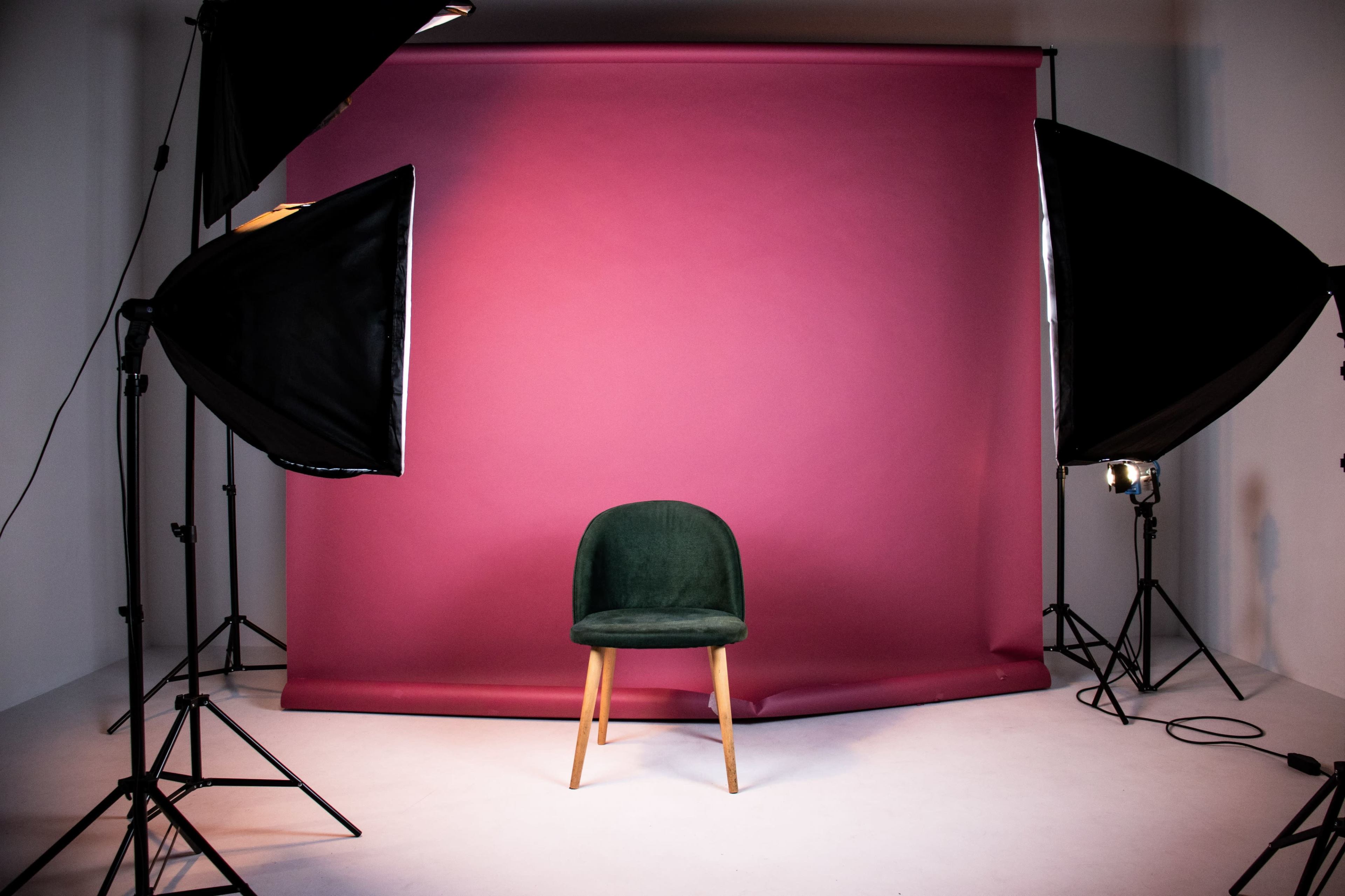 A green chair is positioned in front of a pink backdrop, illuminated by studio lighting equipment.