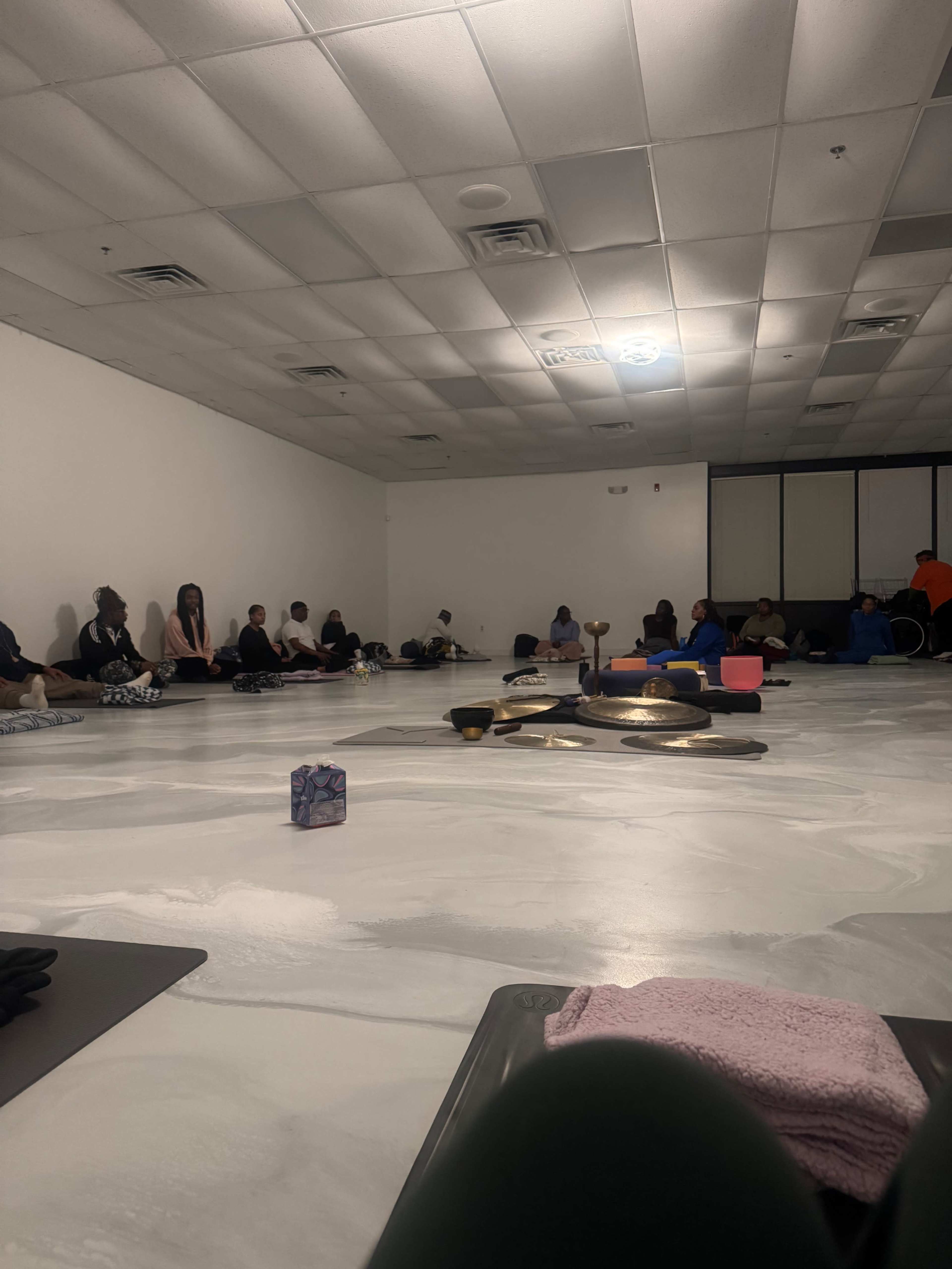 A group of people sits quietly on the floor of a spacious, dimly lit room, with various meditation tools placed in the center.
