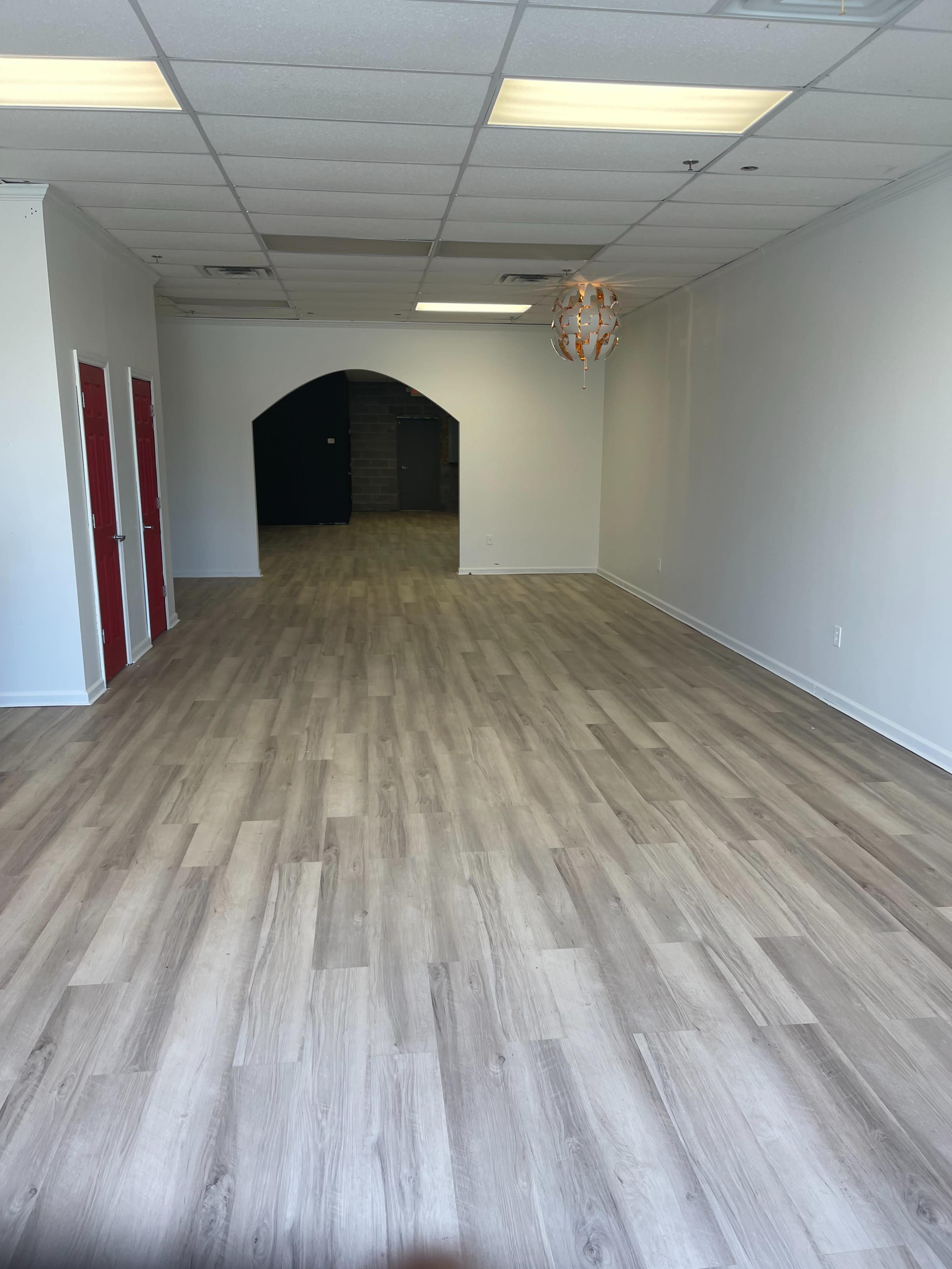 An empty commercial space with light wood flooring and white walls, featuring red doors on one side and an archway leading to a darker area in the back.