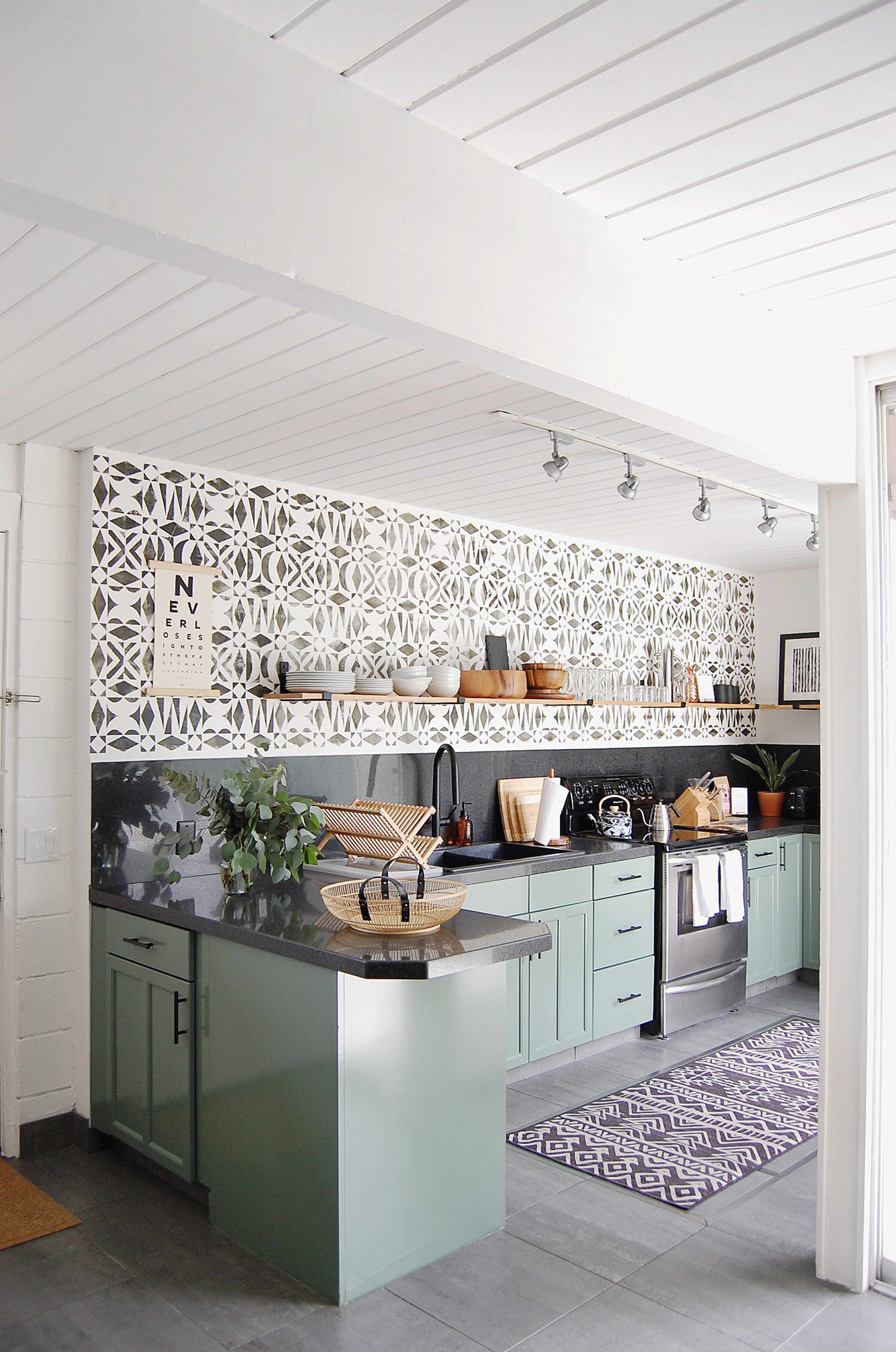 The image shows a modern kitchen with green cabinets, a patterned backsplash, open shelving with kitchenware, and gray floor tiles.