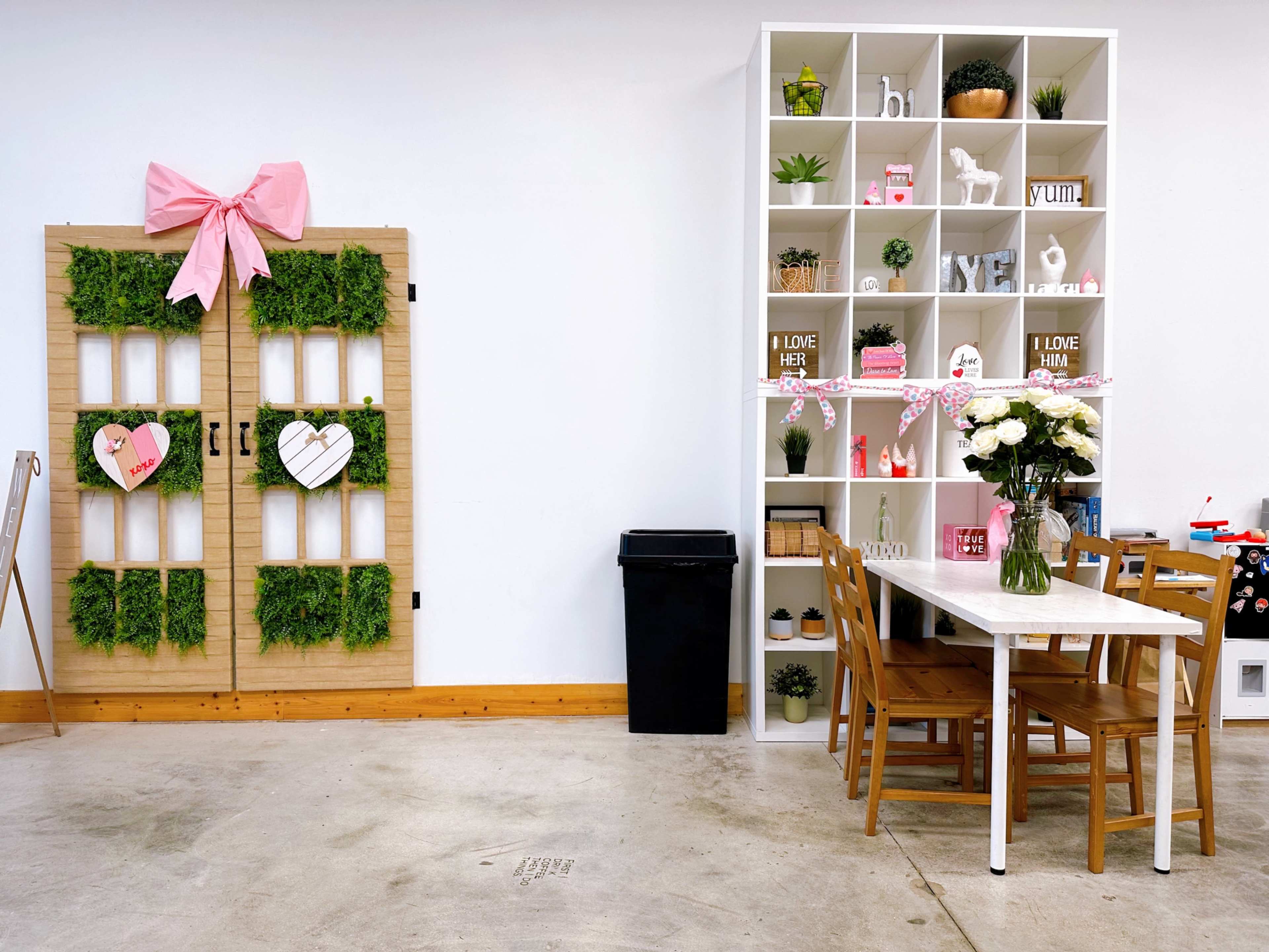 The image shows a bright interior featuring a wooden door with greenery and a pink bow on the left, alongside a white shelf filled with decorative items and a small dining table with chairs on the right.