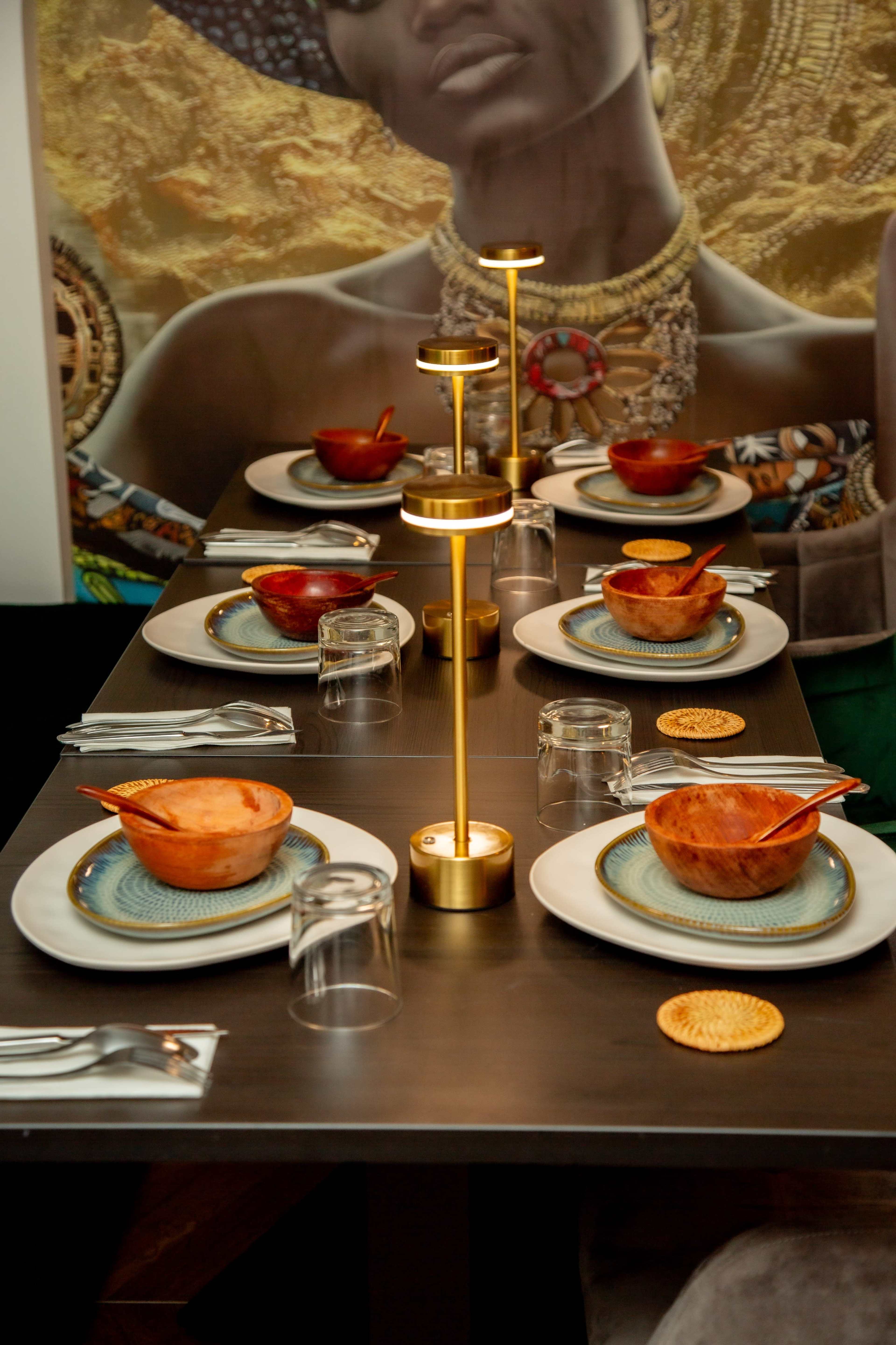 A dining table is set with bowls, plates, and glasses, illuminated by decorative lamps, against a wall featuring an artistic mural.