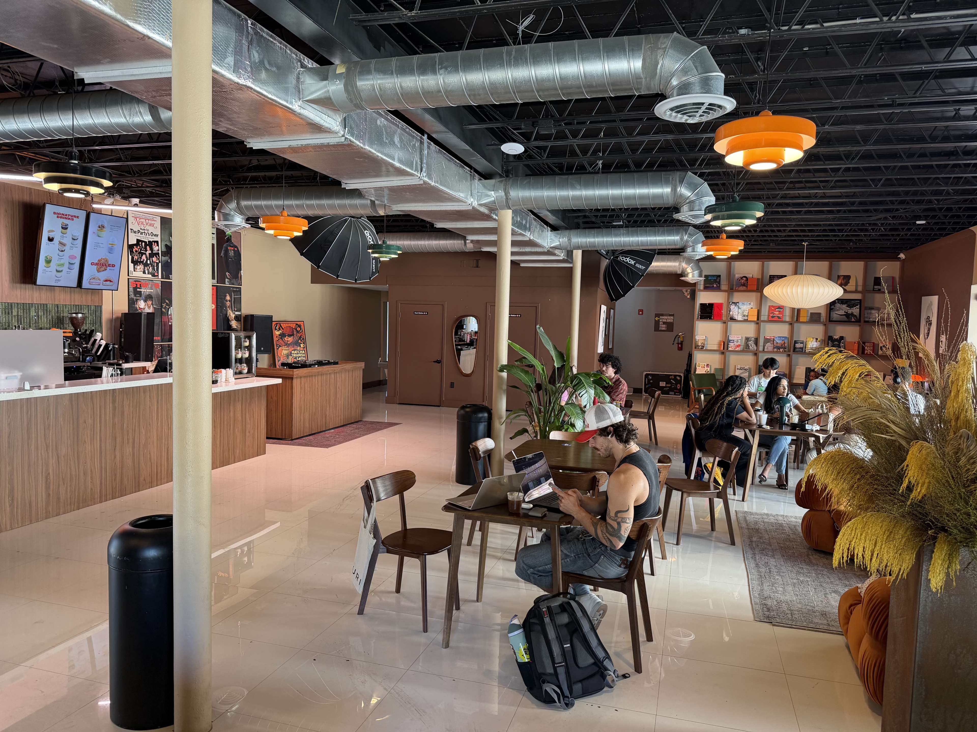 A modern cafe features a spacious interior with a seating area, plants, and various lighting fixtures, filled with customers engaged in activities like working and socializing.