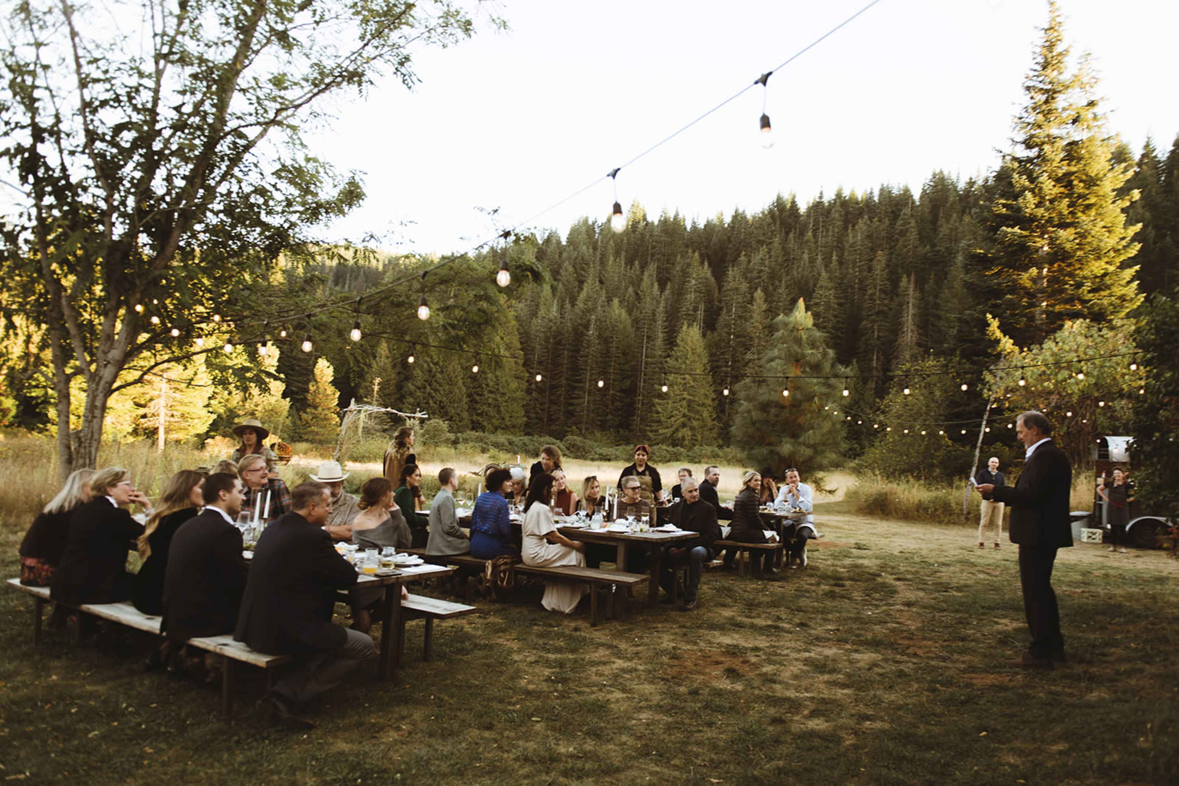 A group of people is seated at long picnic tables in a picturesque outdoor setting with string lights and trees in the background.