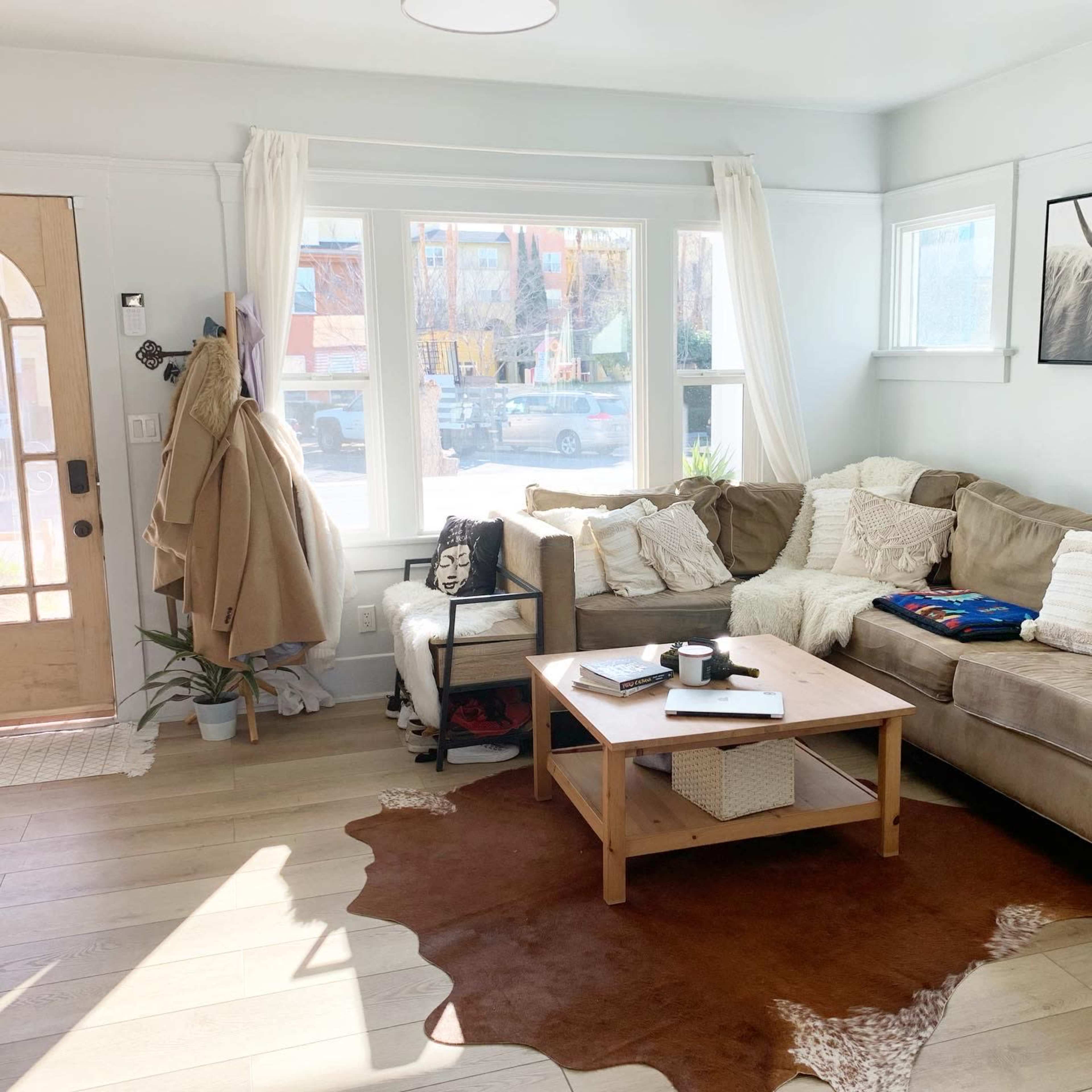 A living room features a beige sectional couch, a wooden coffee table, and large windows allowing natural light to enter, with decor including a rug and potted plant.