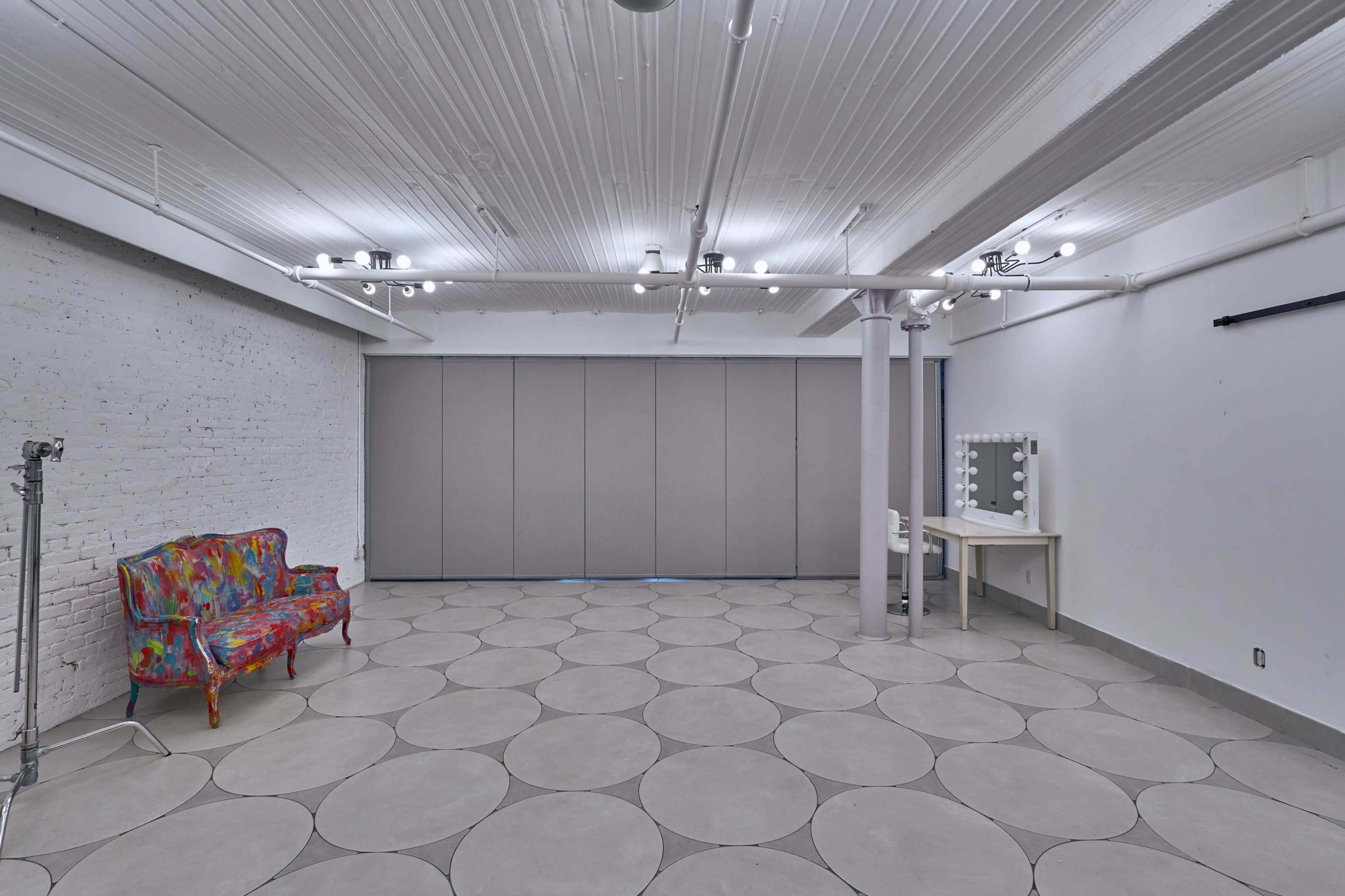 A spacious interior room with a patterned floor, a colorful sofa, a makeup vanity with lights, and gray folding doors.