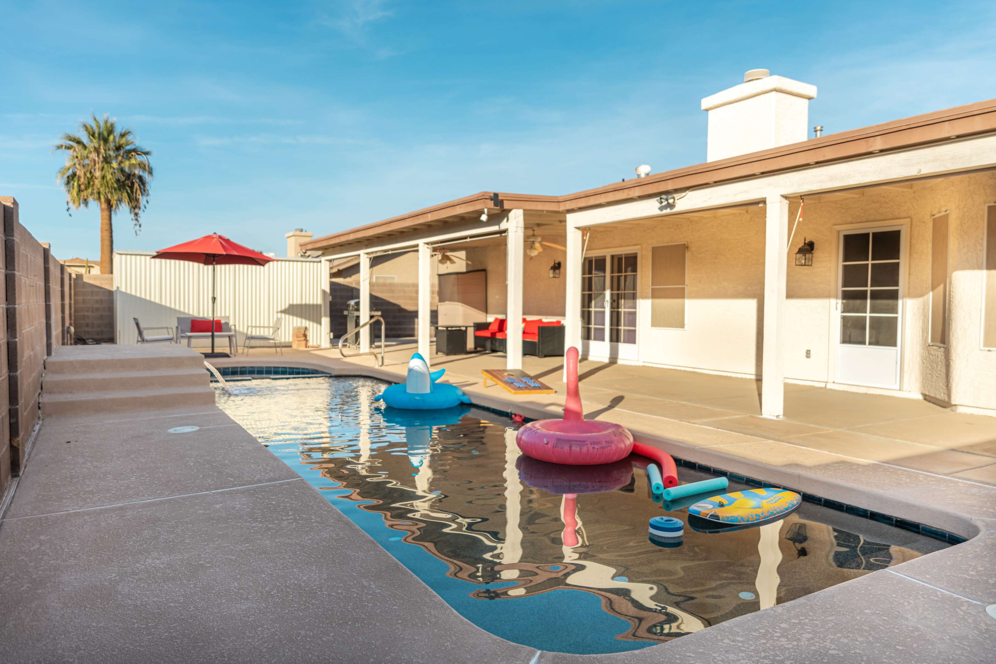 A residential backyard with a swimming pool featuring various inflatable floaties and a patio area with an umbrella.