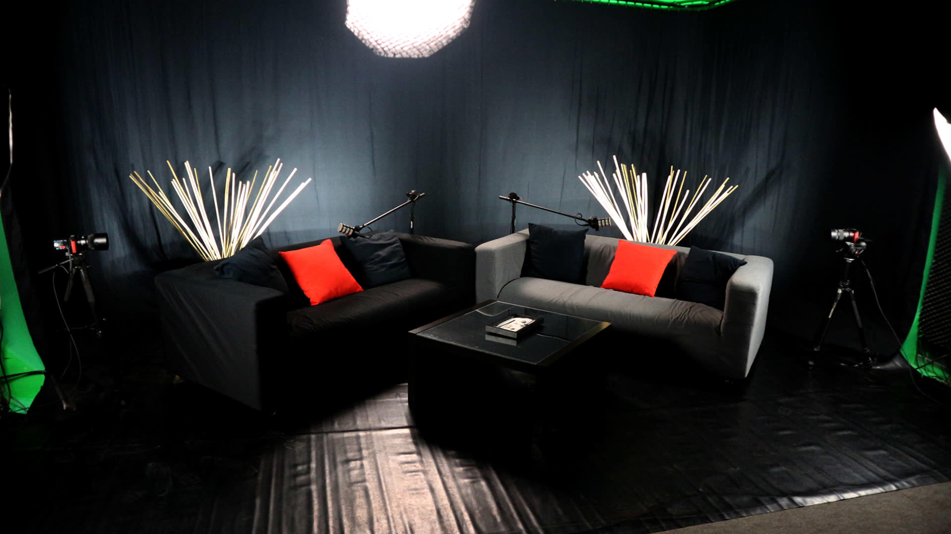 The image shows two sofas—one black and one gray—arranged facing each other with red and black cushions, set in a studio environment with cameras and lighting equipment.
