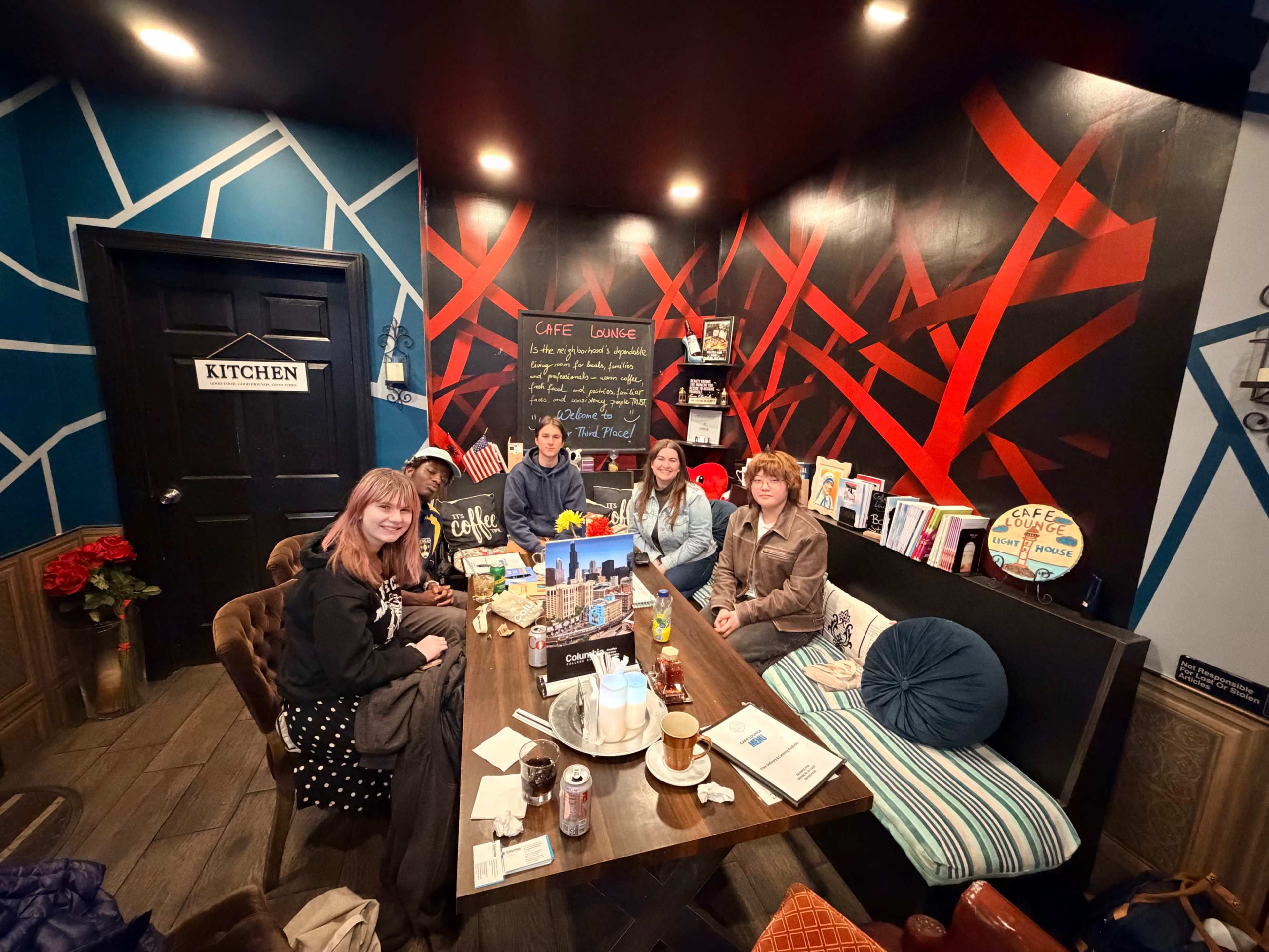 A group of five people is seated at a table in a cozy café with vibrant wall decorations and various items displayed around them.