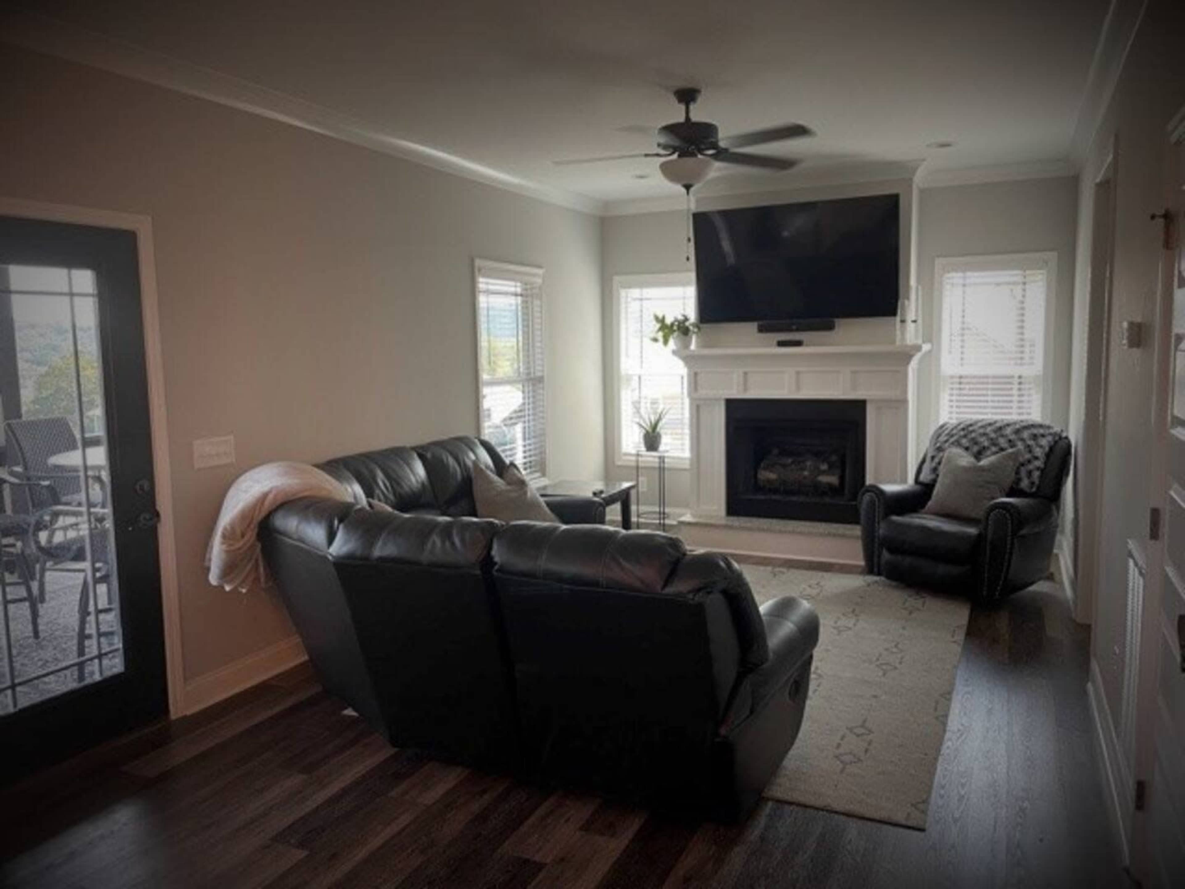 A living room with a black leather sofa, an armchair, a television mounted above a white fireplace, and large windows allowing natural light.