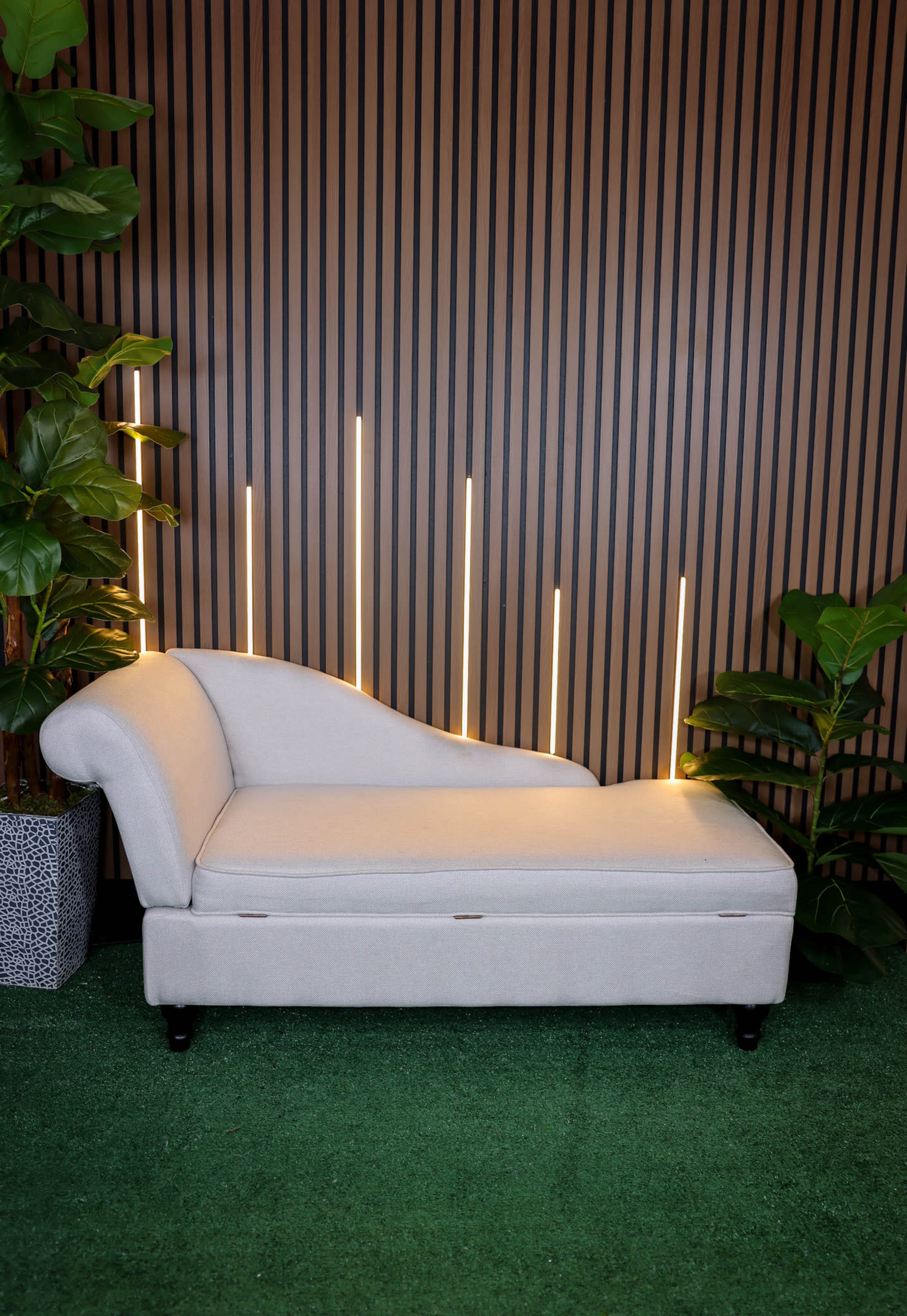 A light-colored chaise lounge is positioned against a backdrop of vertical wooden slats, with subtle lighting and potted plants on either side.
