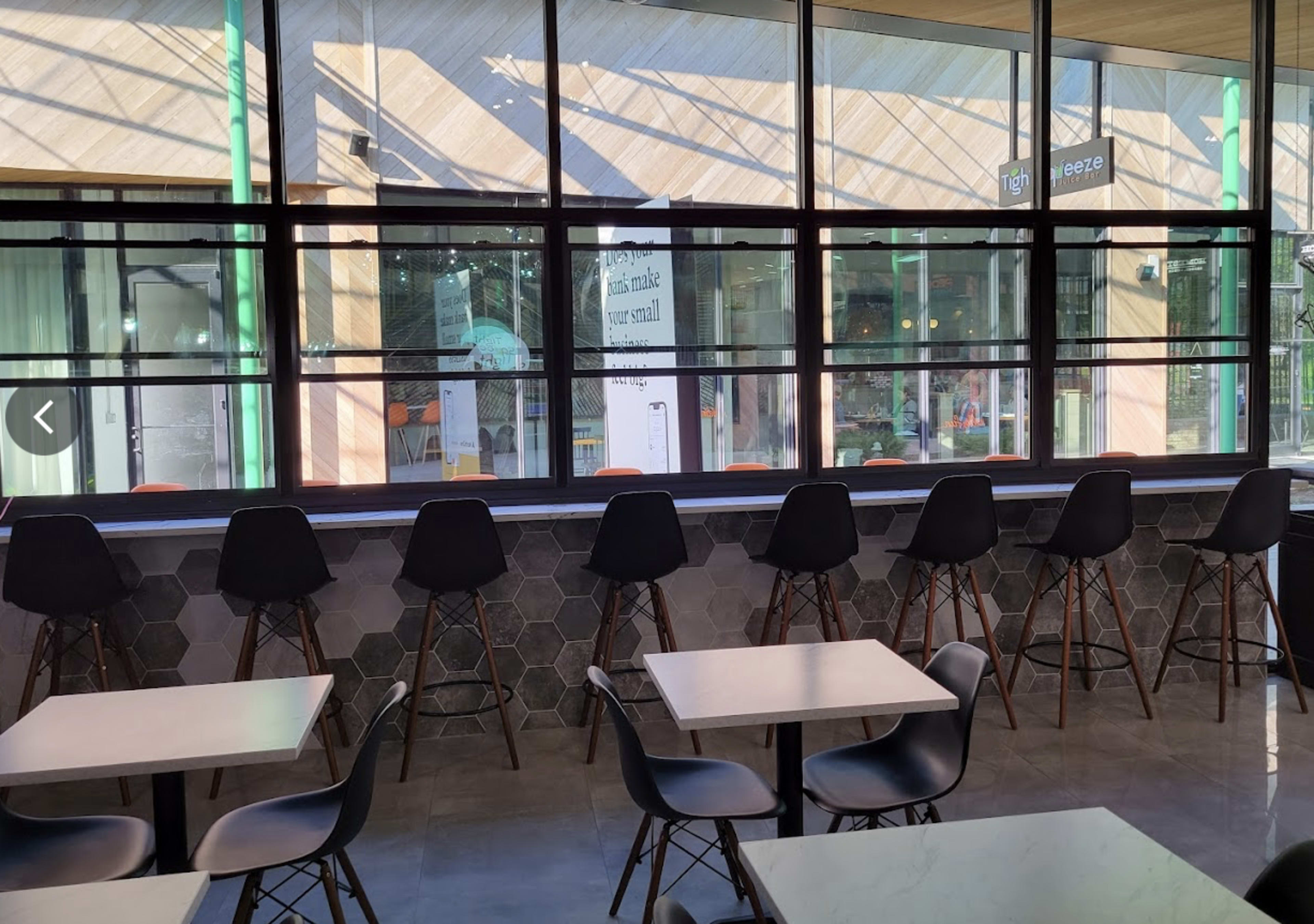 The image shows a modern cafe interior with a row of tall black stools facing a large window, surrounded by tables and a patterned wall.