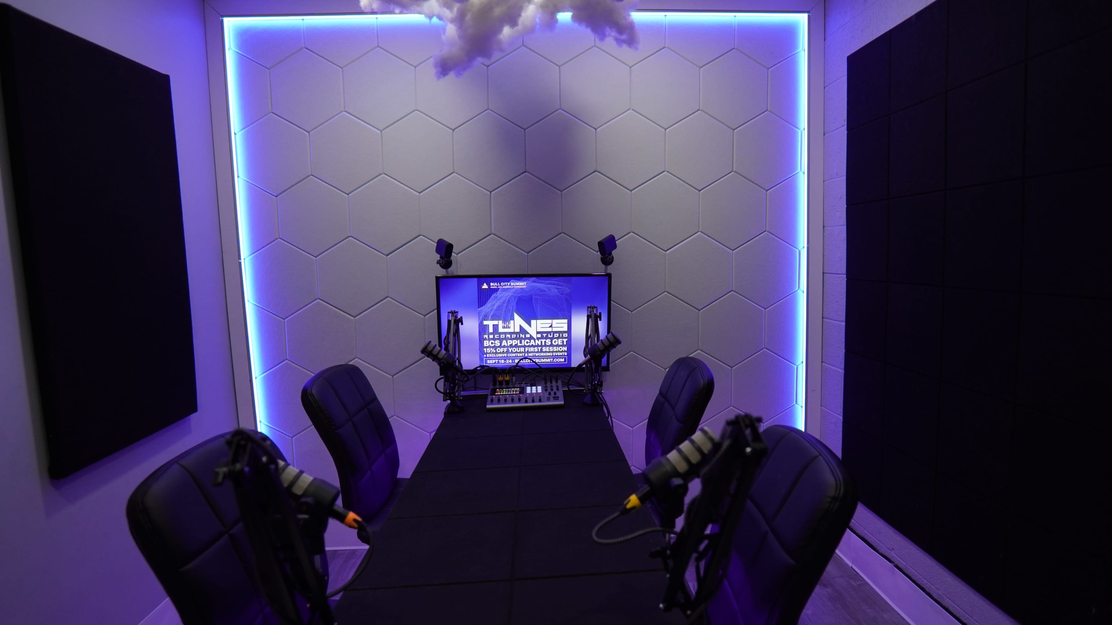 The image shows a modern recording studio featuring hexagon-patterned wall panels, a central table with four chairs, a display screen, and microphone setups under soft lighting.