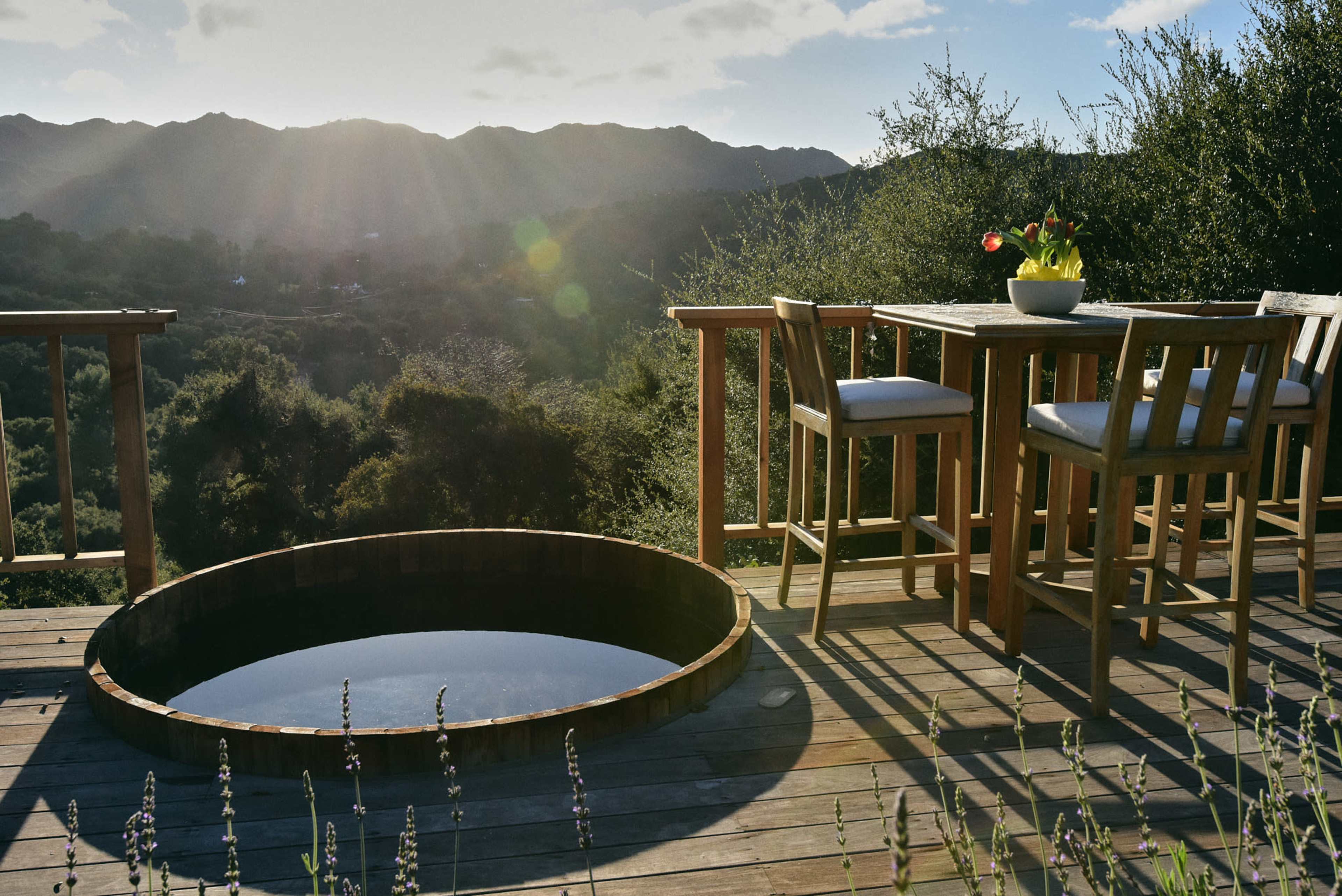 A wooden deck features a round hot tub and a dining table set against a backdrop of mountains and a setting sun.