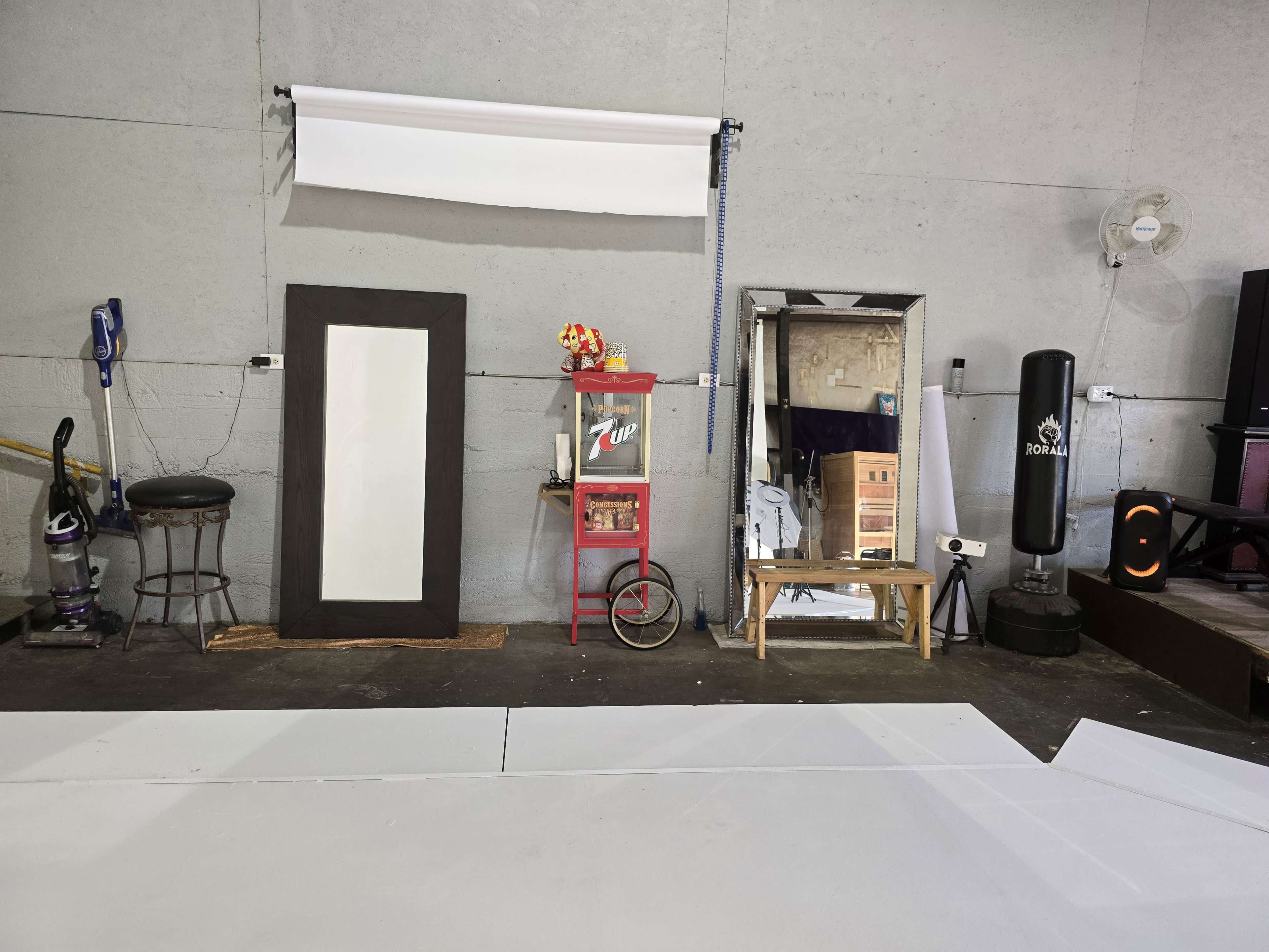 The image depicts a room with a gray wall featuring various items, including mirrors, a stool, a vacuum cleaner, a boxing bag, and a vintage soda display.
