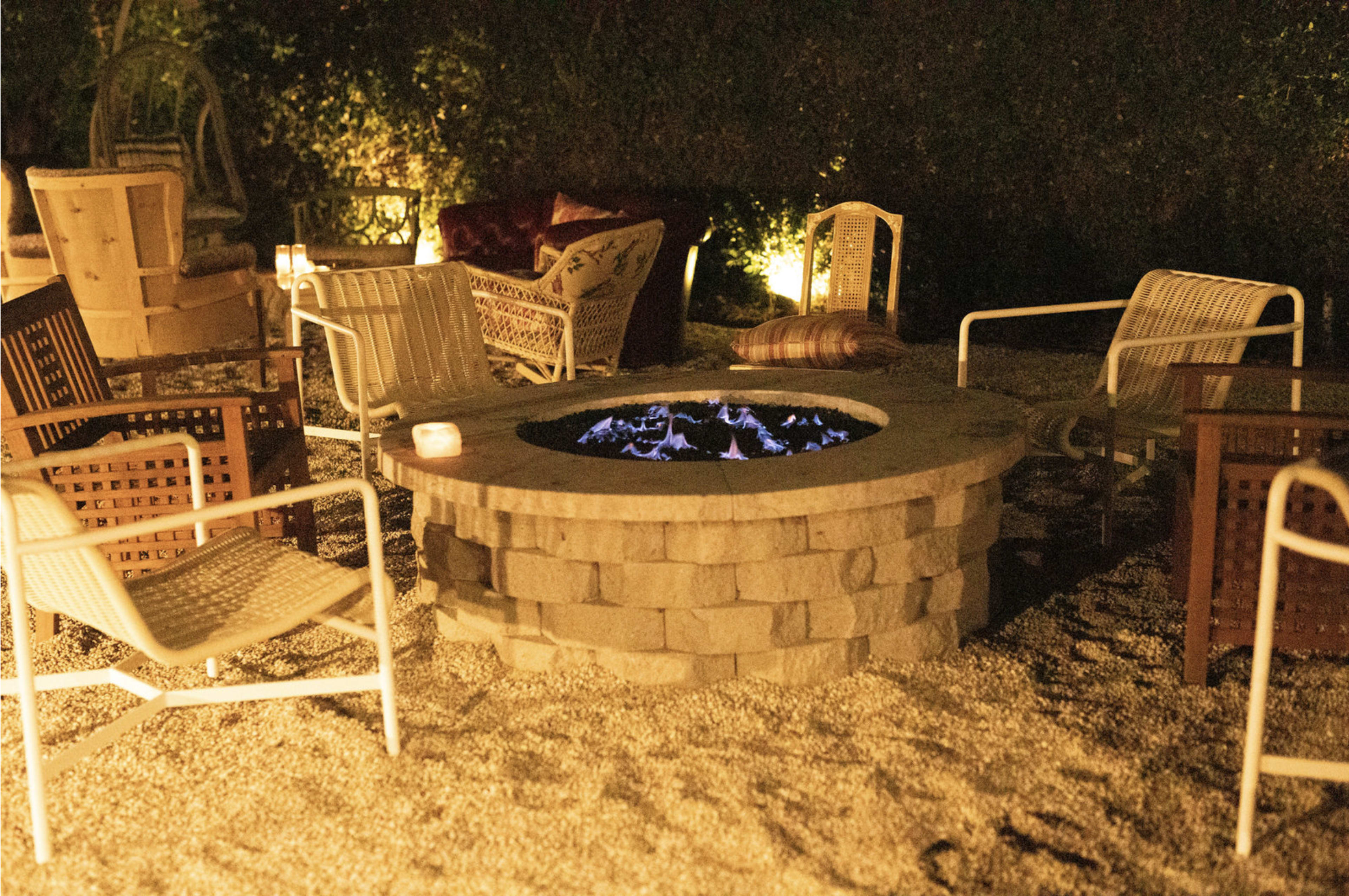 The image shows a circular stone fire pit surrounded by several chairs in a dimly lit outdoor setting.