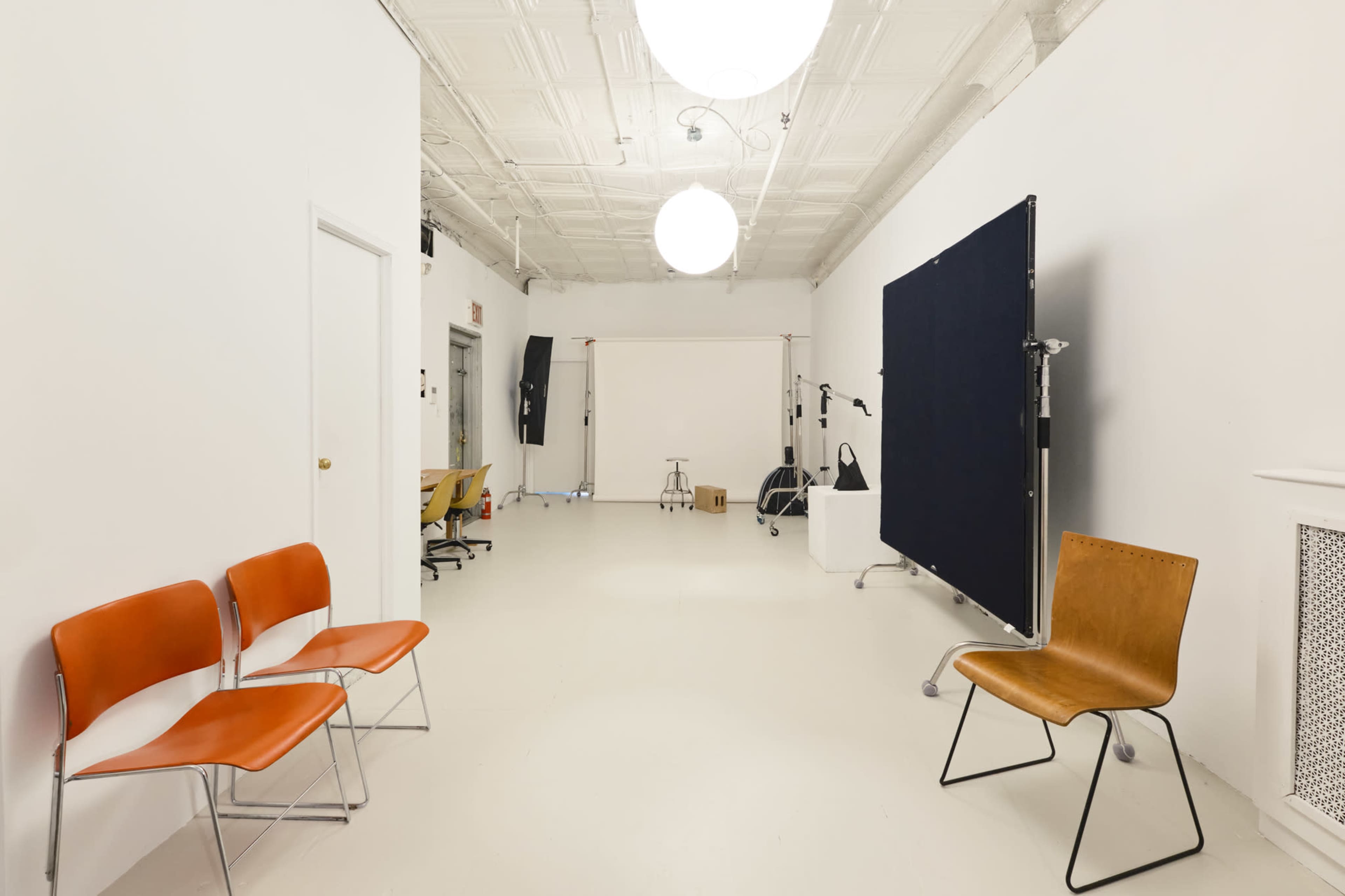 The image shows a minimalist studio space with white walls, a light-colored floor, and a few chairs arranged against one side.