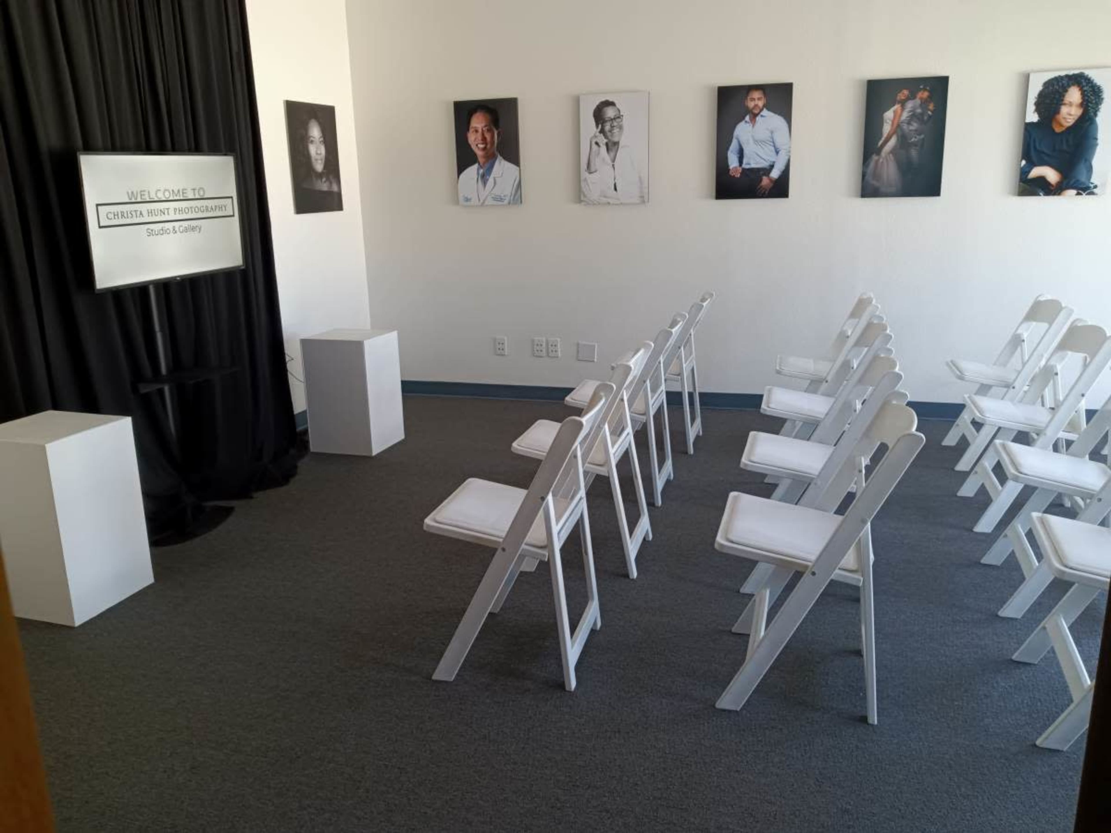 The image depicts a gallery space with white chairs arranged in rows facing a backdrop featuring portraits on the wall.