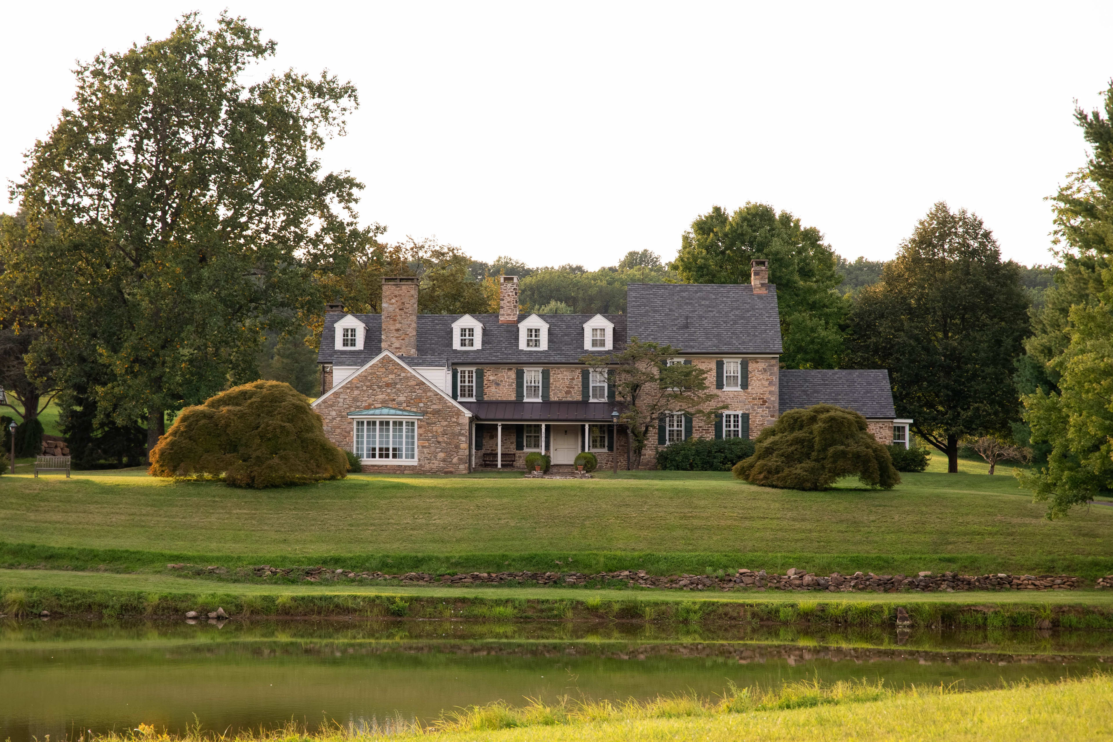 The image shows a large, stone house with multiple stories and a front porch set on a grassy landscape next to a calm body of water.