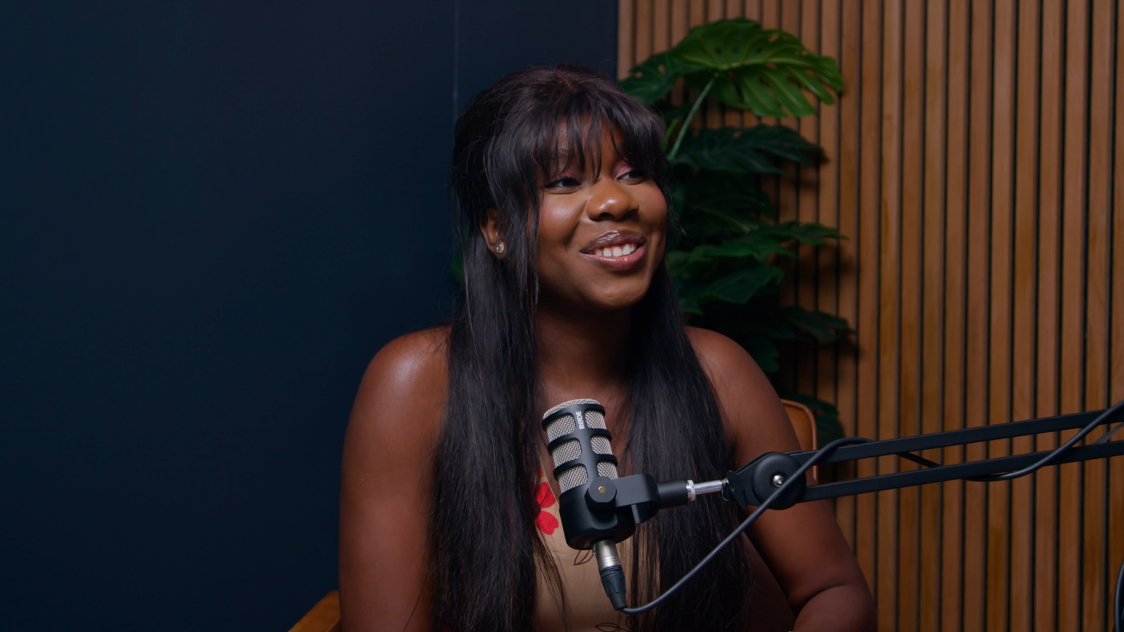 A woman with long dark hair sits in front of a microphone, smiling in a well-lit room with wooden paneling and a plant in the background.