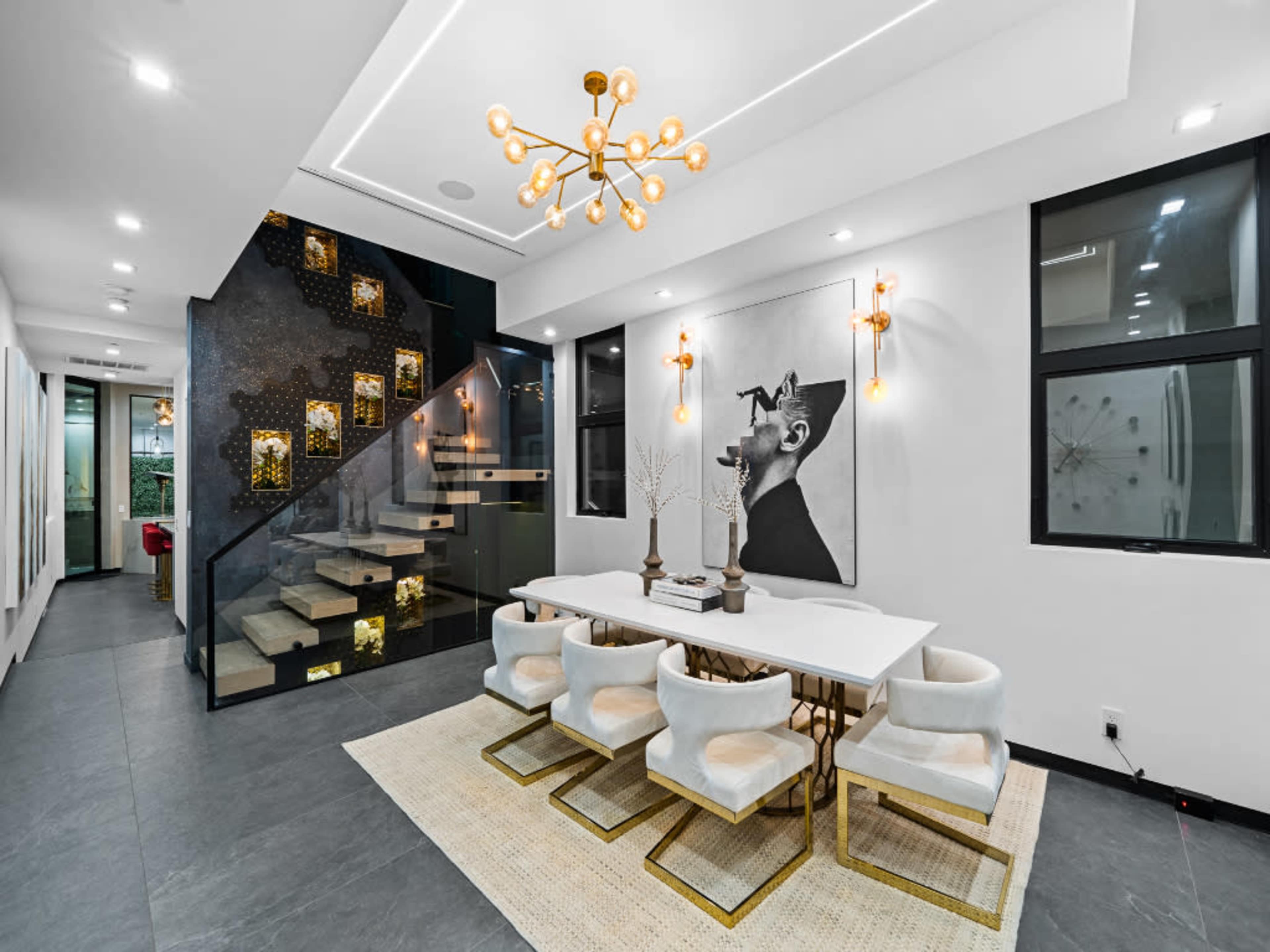 The image shows a modern dining area featuring a white table surrounded by upholstered chairs, with a staircase in the background that has decorative lighting and a large art piece on the wall.