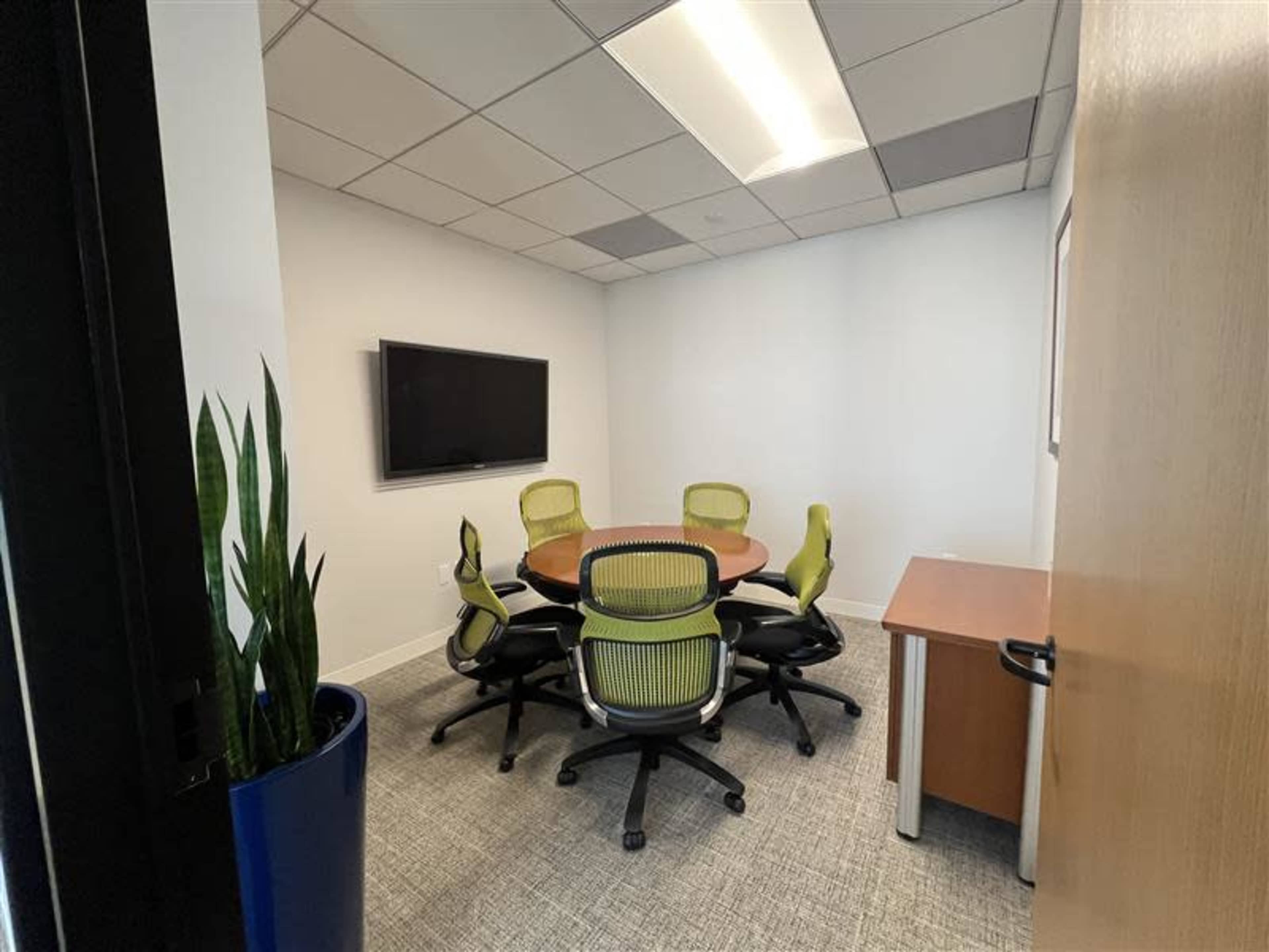 The image shows a small conference room with a round table, six green office chairs, a television on the wall, and a potted plant in the corner.