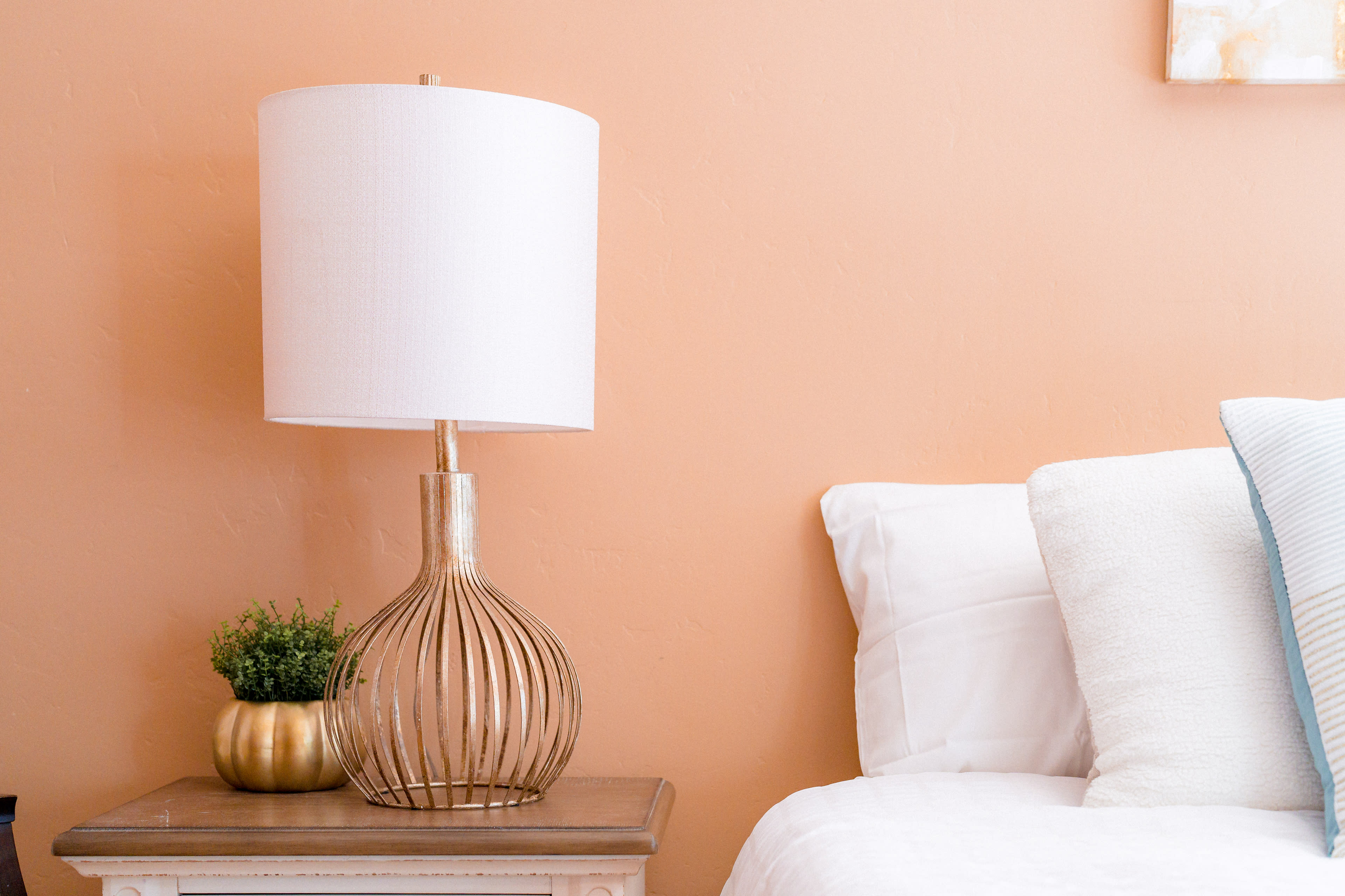 A lamp with a white shade is placed on a wooden side table next to a neatly made bed against a peach-colored wall.