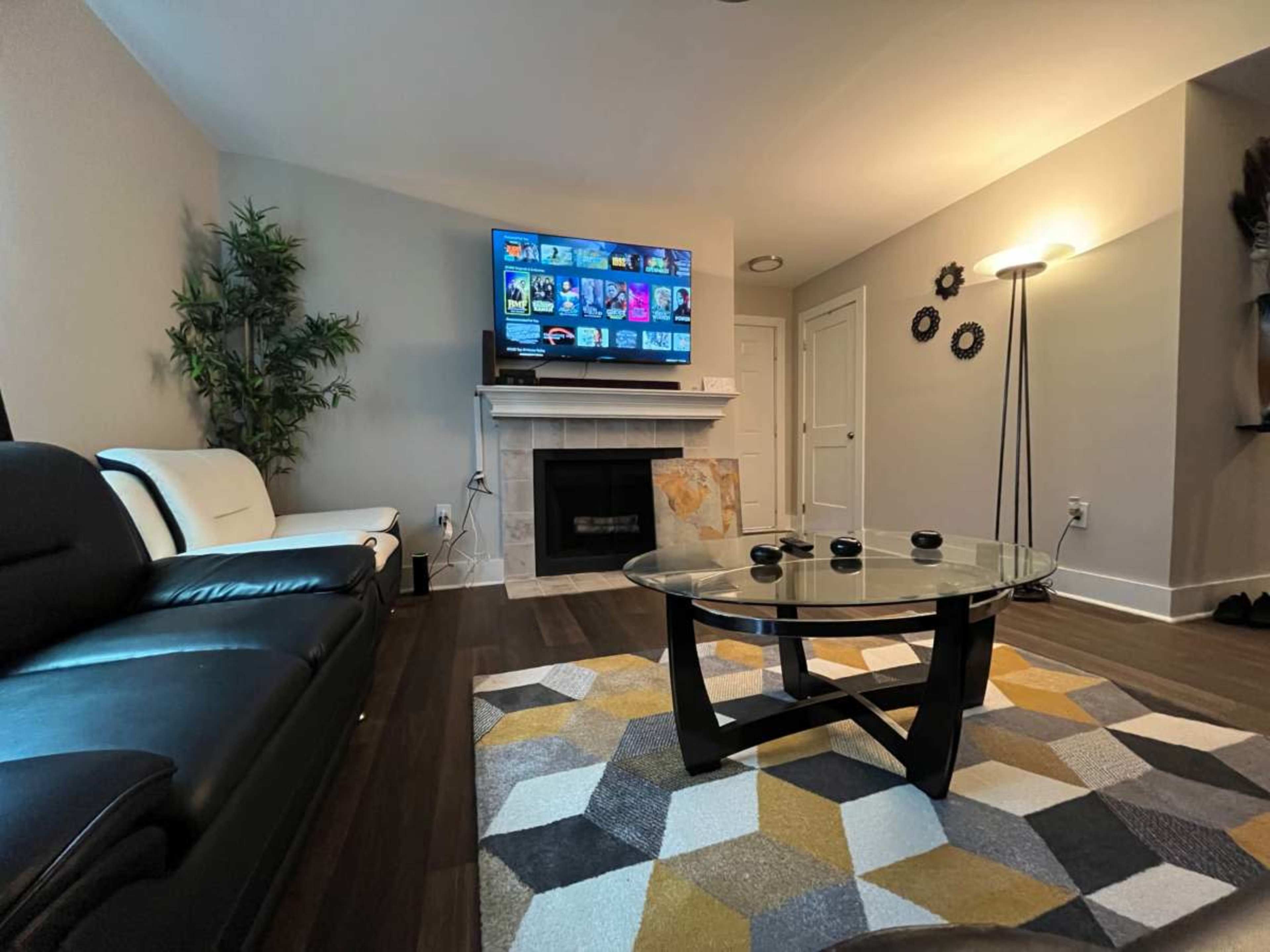 The image shows a modern living room with a black and white couch, a glass coffee table on a patterned rug, and a mounted television above a fireplace.