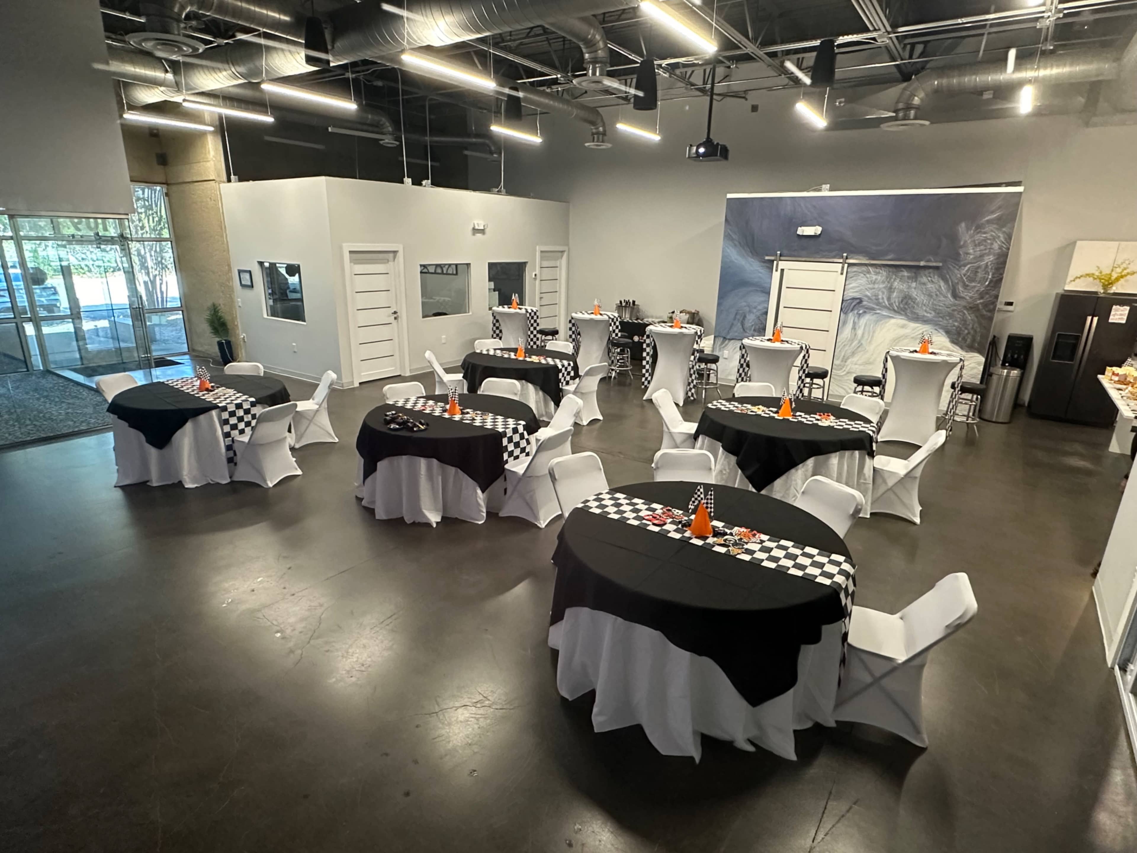 The image shows a large event space arranged with several round tables covered in black and white tablecloths, decorated with centerpieces, and set with chairs.
