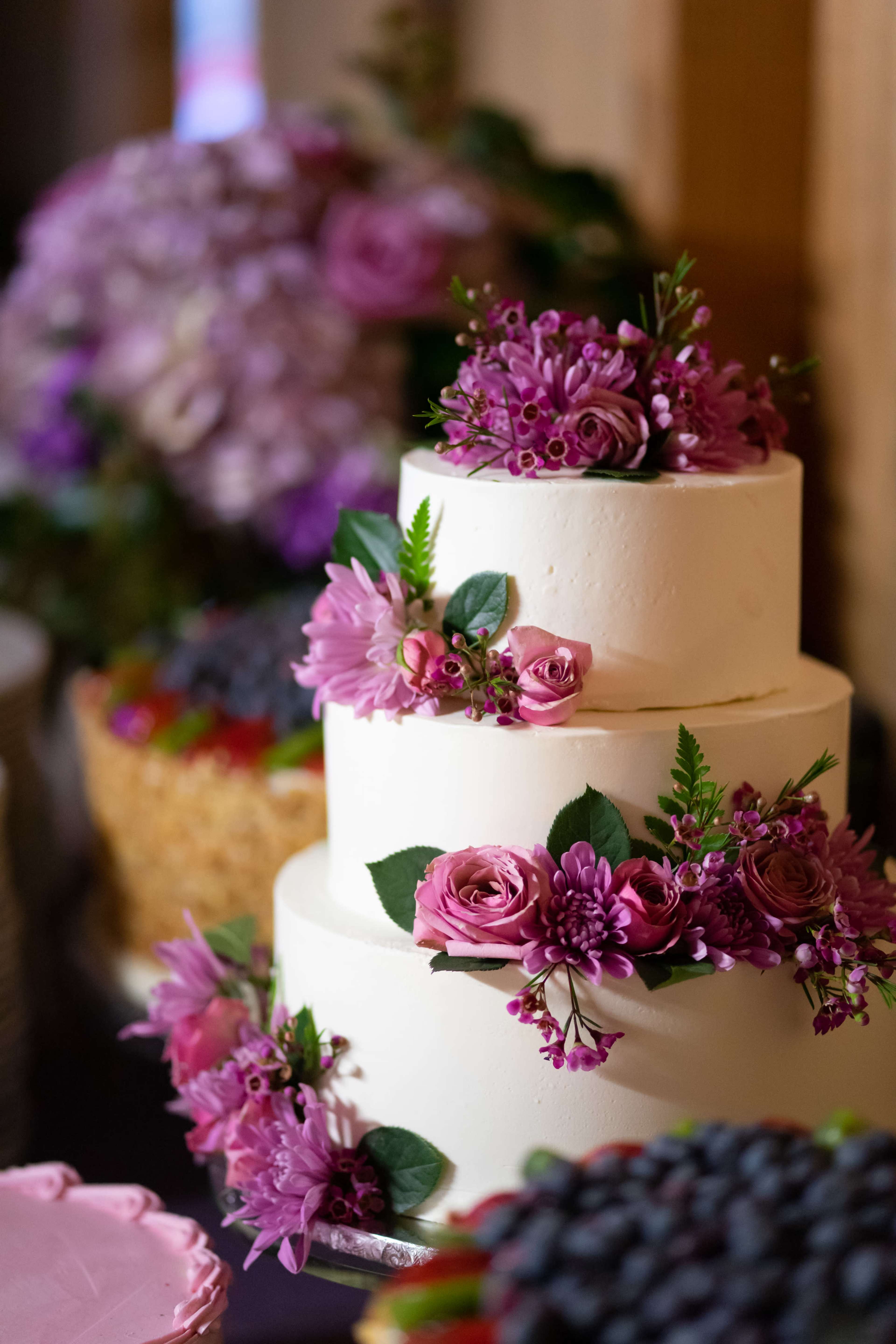 A three-tiered white wedding cake is decorated with various pink flowers and greenery, positioned alongside other desserts.