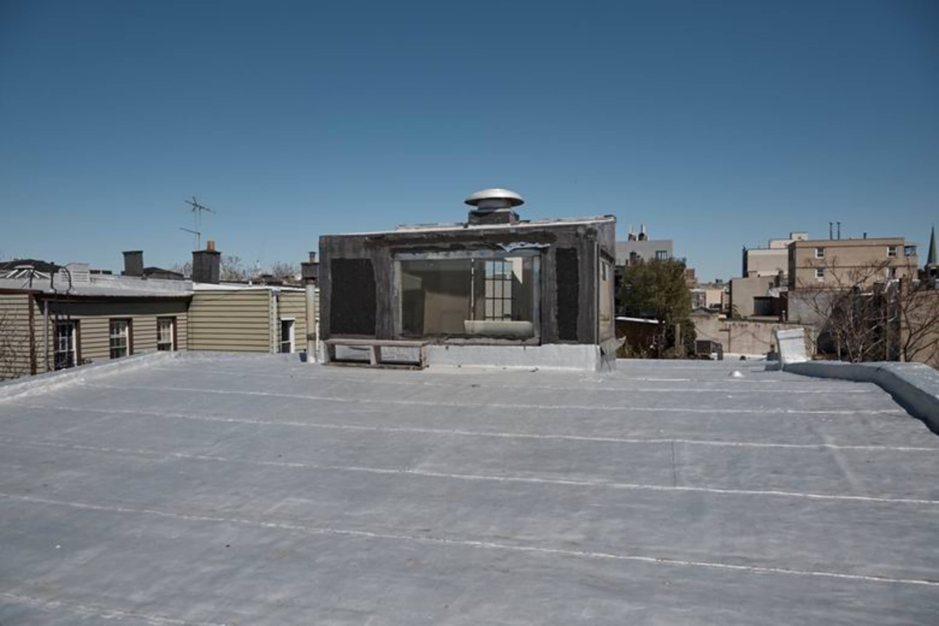 The image shows a flat rooftop with a skylight structure and nearby buildings under a clear blue sky.
