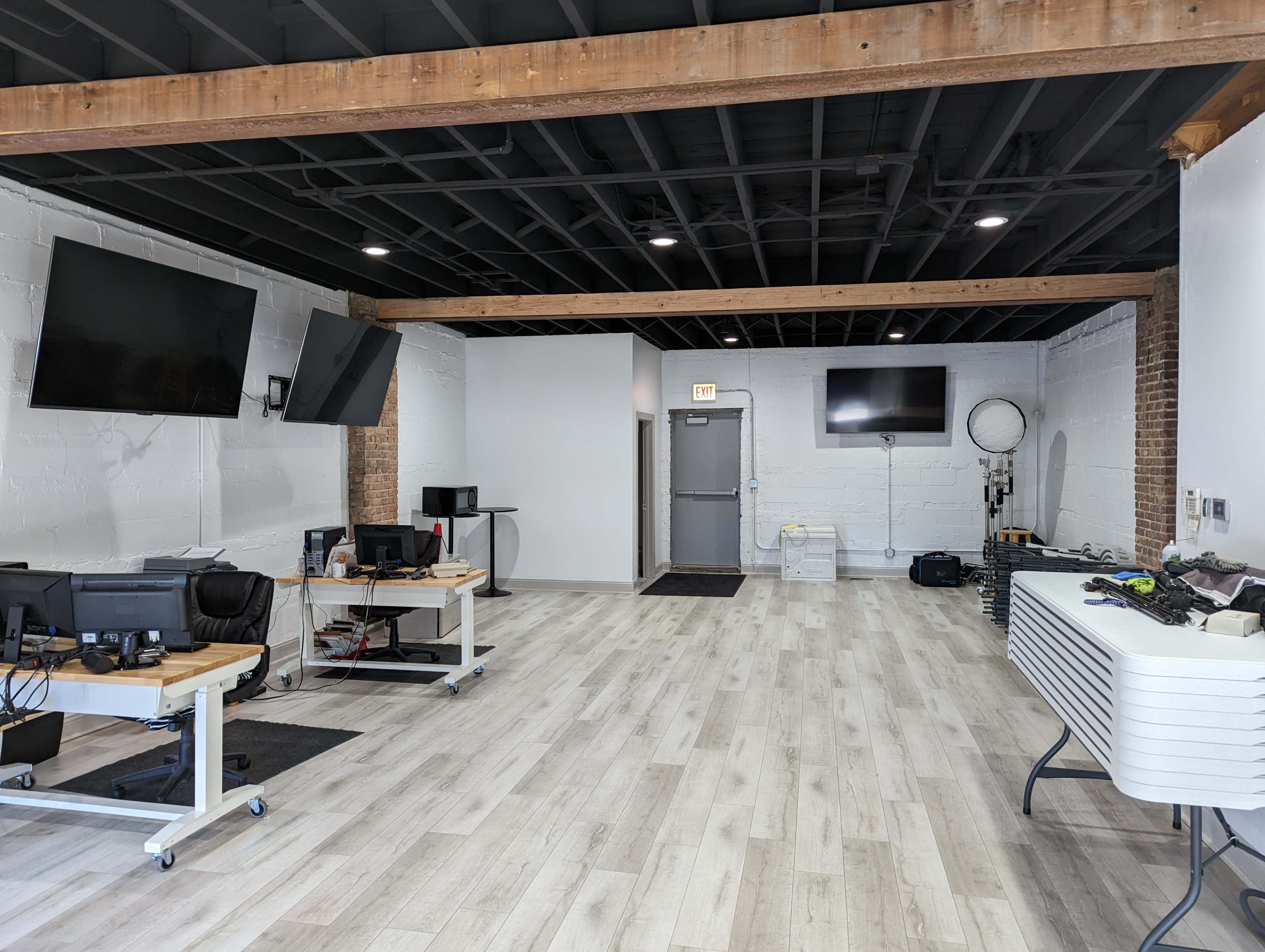 An open office space with multiple workstations, two large wall-mounted screens, and a door leading to another area.