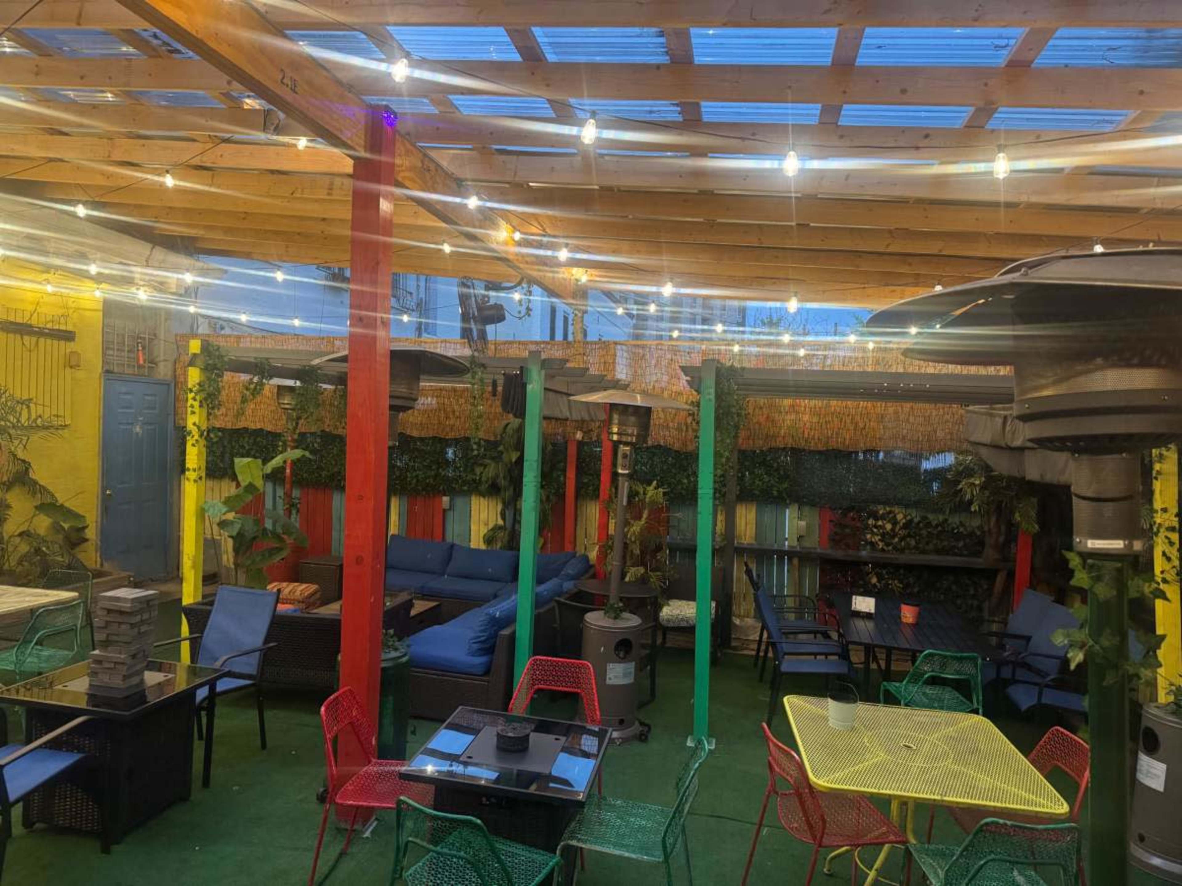 A colorful outdoor dining area features various tables and chairs, string lights overhead, and heating lamps in a partially enclosed space.