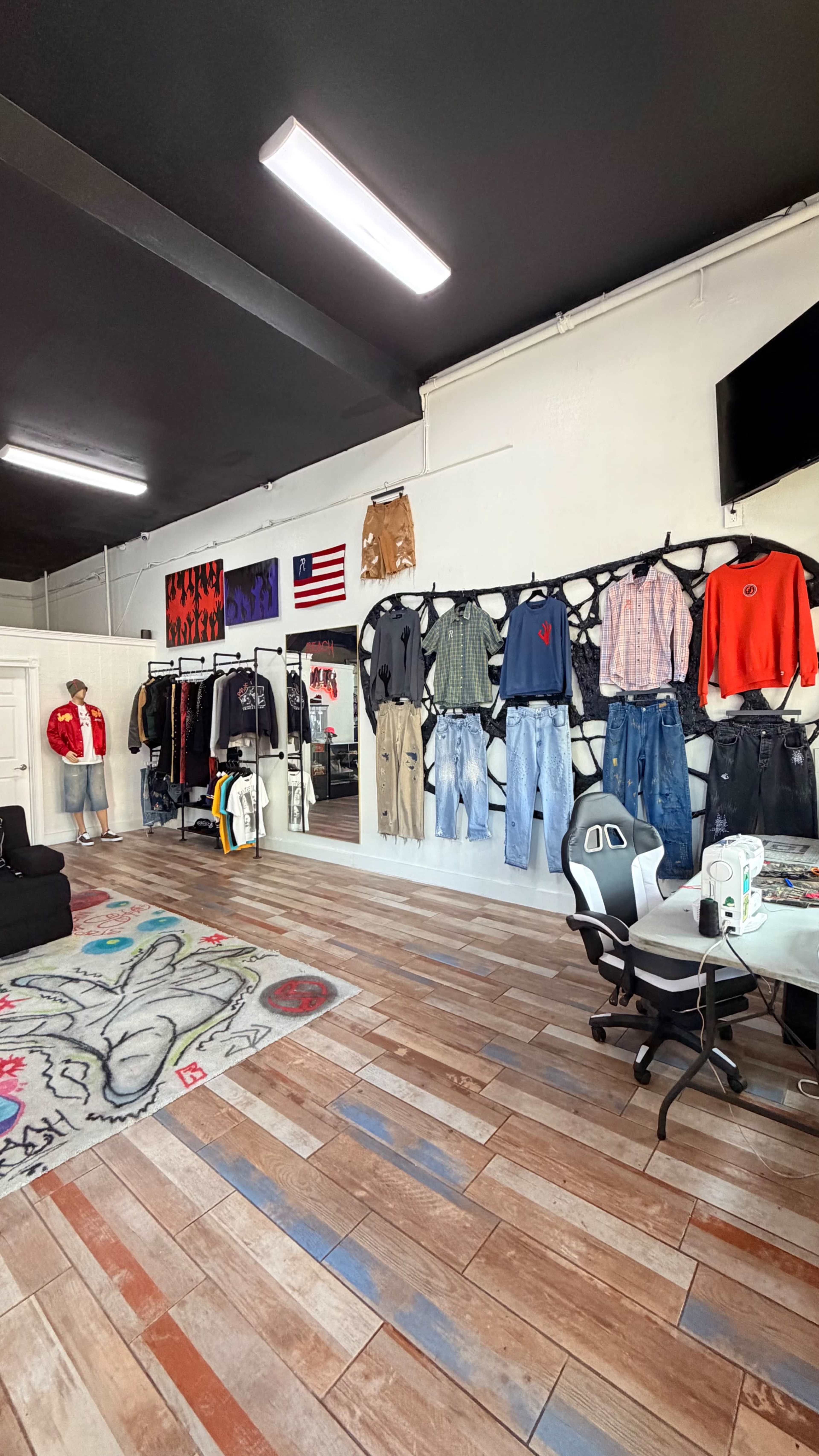 The image shows a clothing shop interior with various garments displayed on a wall and a seating area featuring a table and chairs.