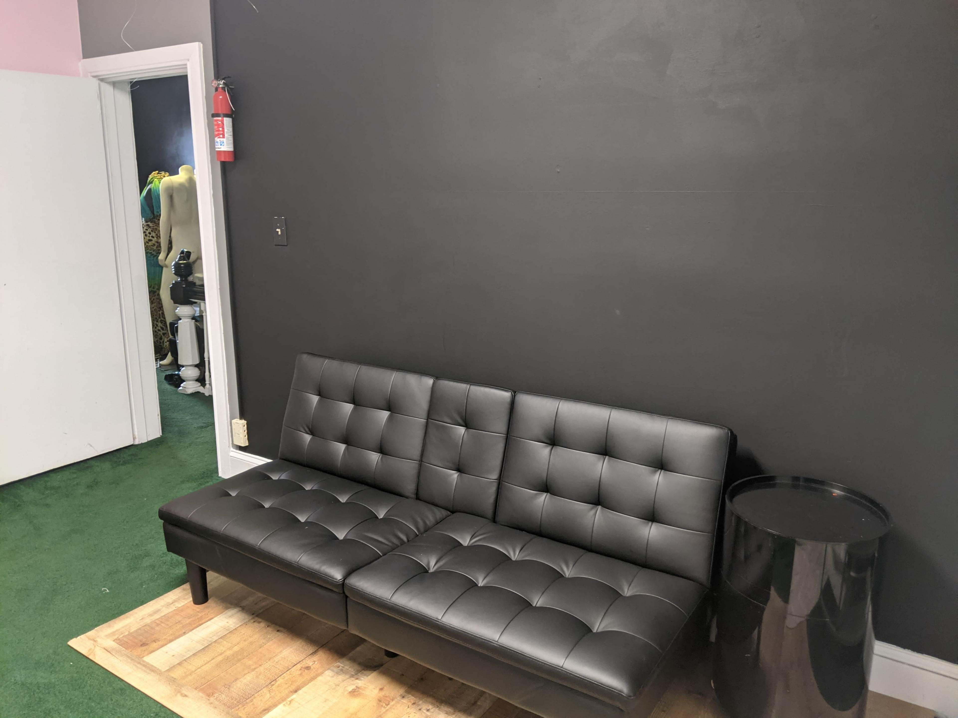 A black futon is placed against a dark wall next to a round black side table in a room with green carpet.