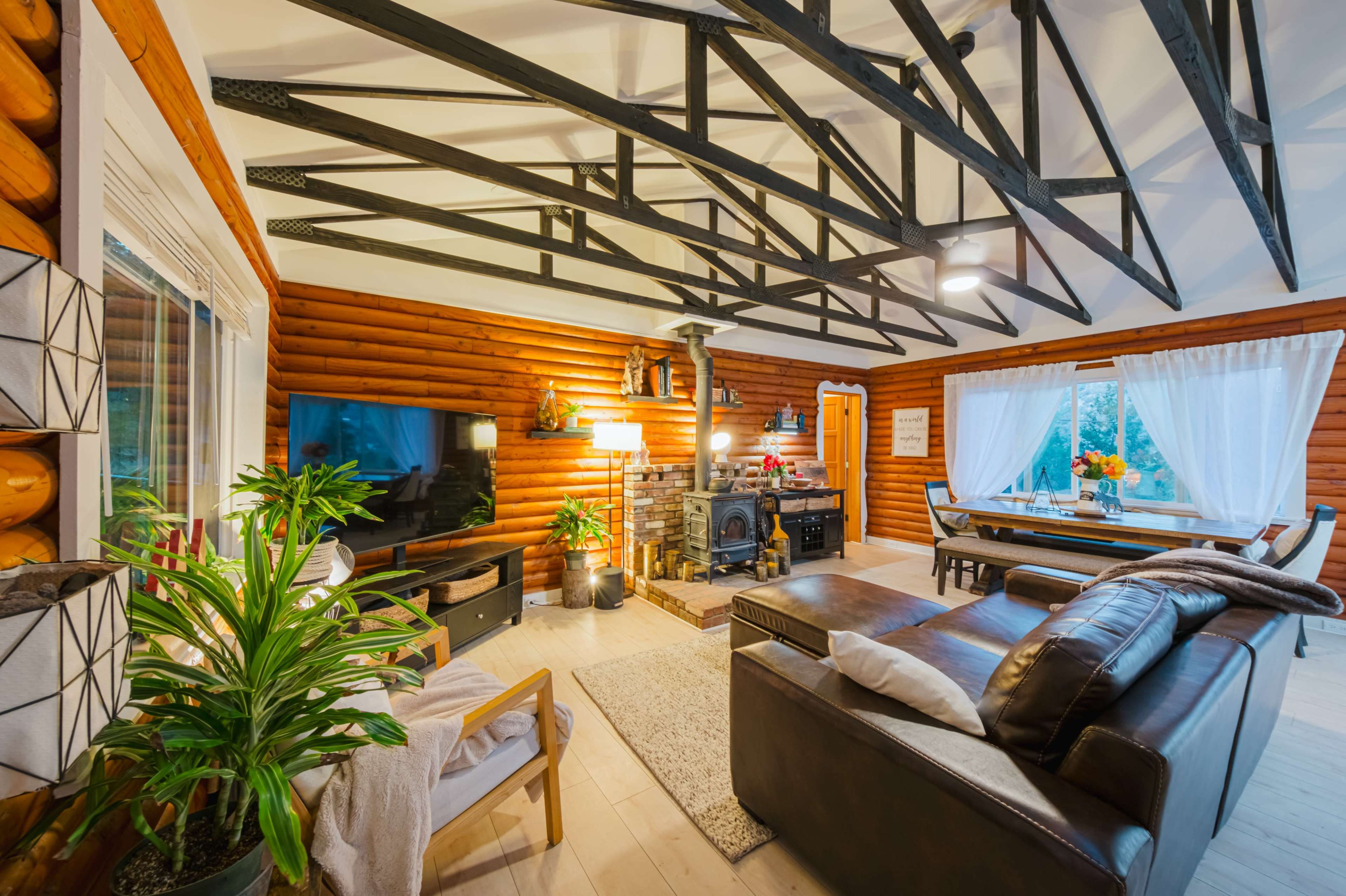 A cozy living room with wooden log walls, a fireplace, a dining area, and large windows letting in natural light.