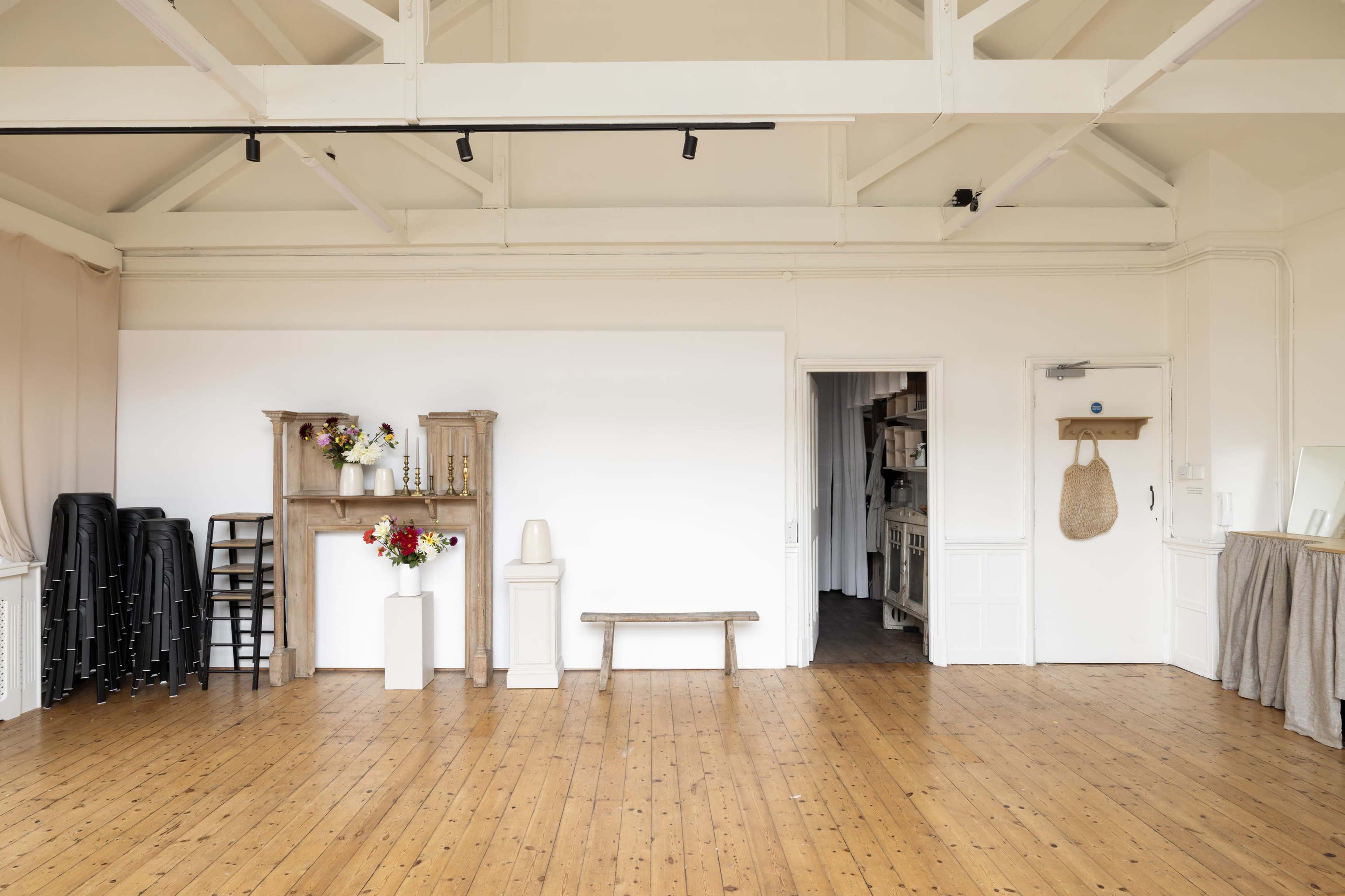 The image shows a spacious, bright room with wooden flooring, featuring a few chairs stacked against one wall, decorative shelves, a flower arrangement, a bench, and a doorway leading to another area.
