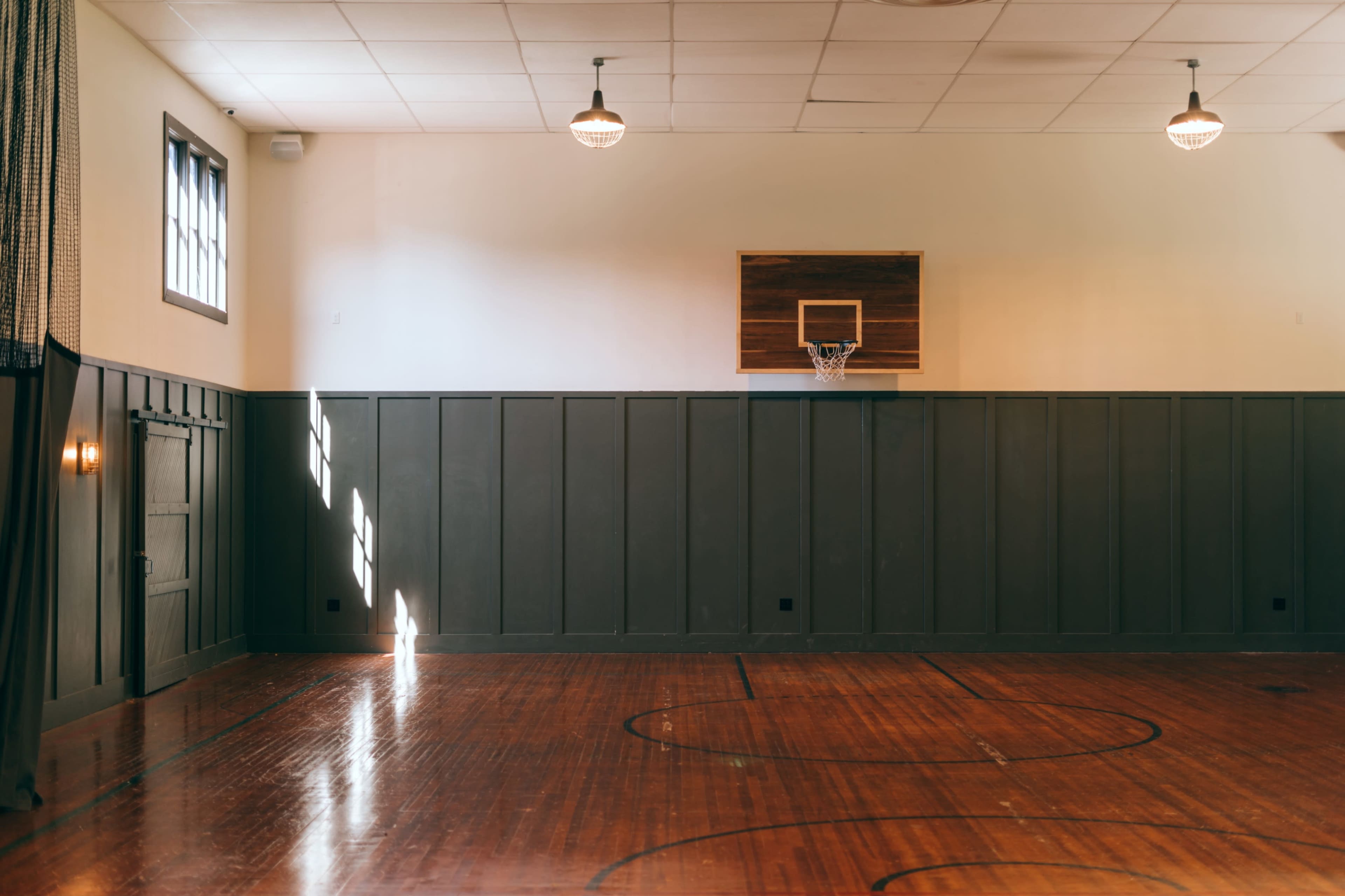 The image shows a gymnasium with a polished wooden floor, a basketball hoop mounted on the wall, and sunlight streaming through the windows.