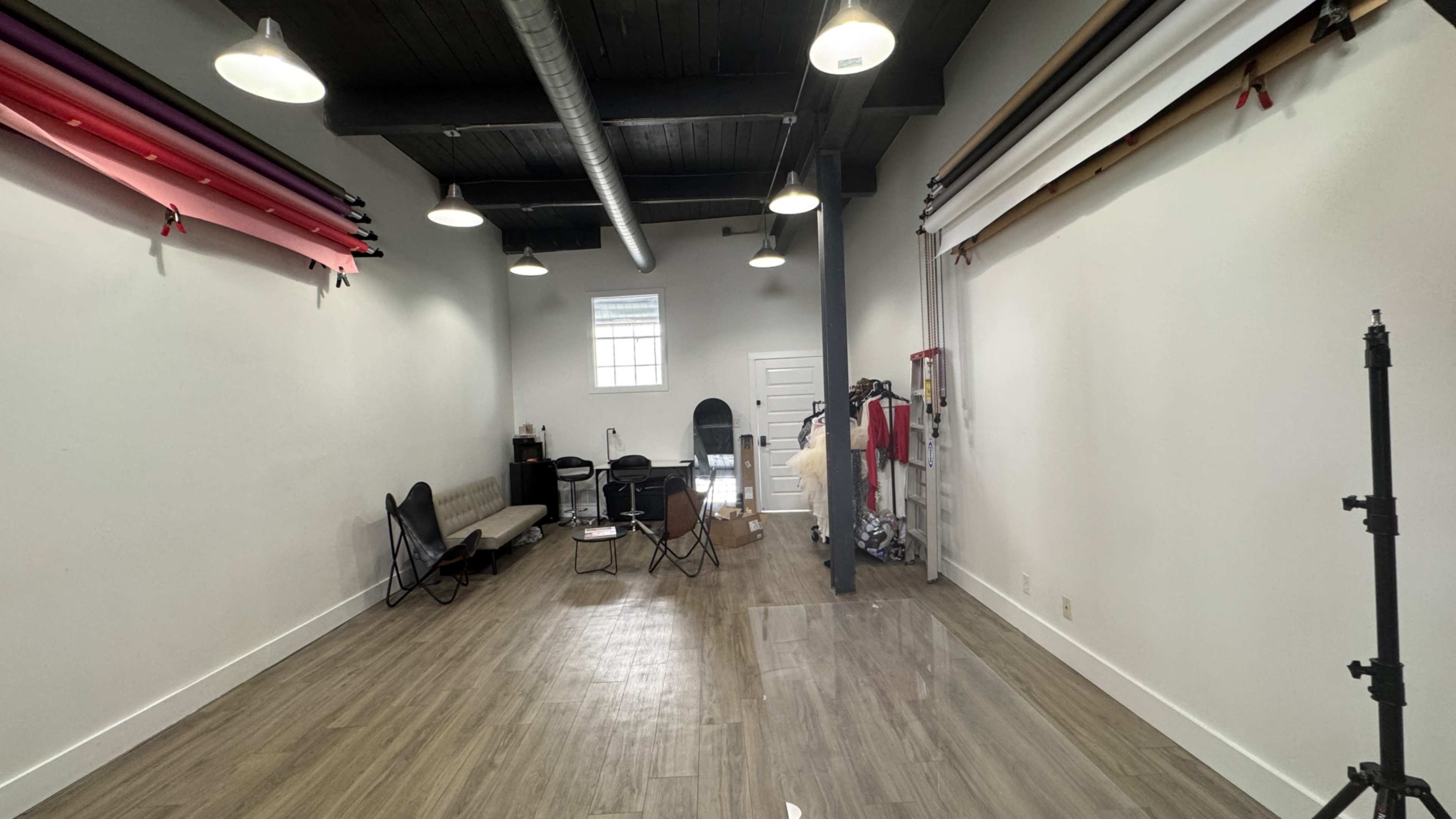 The image shows a spacious, well-lit studio with wooden flooring, equipped with black chairs, a mirror, and colorful backdrop rolls hanging on the walls.