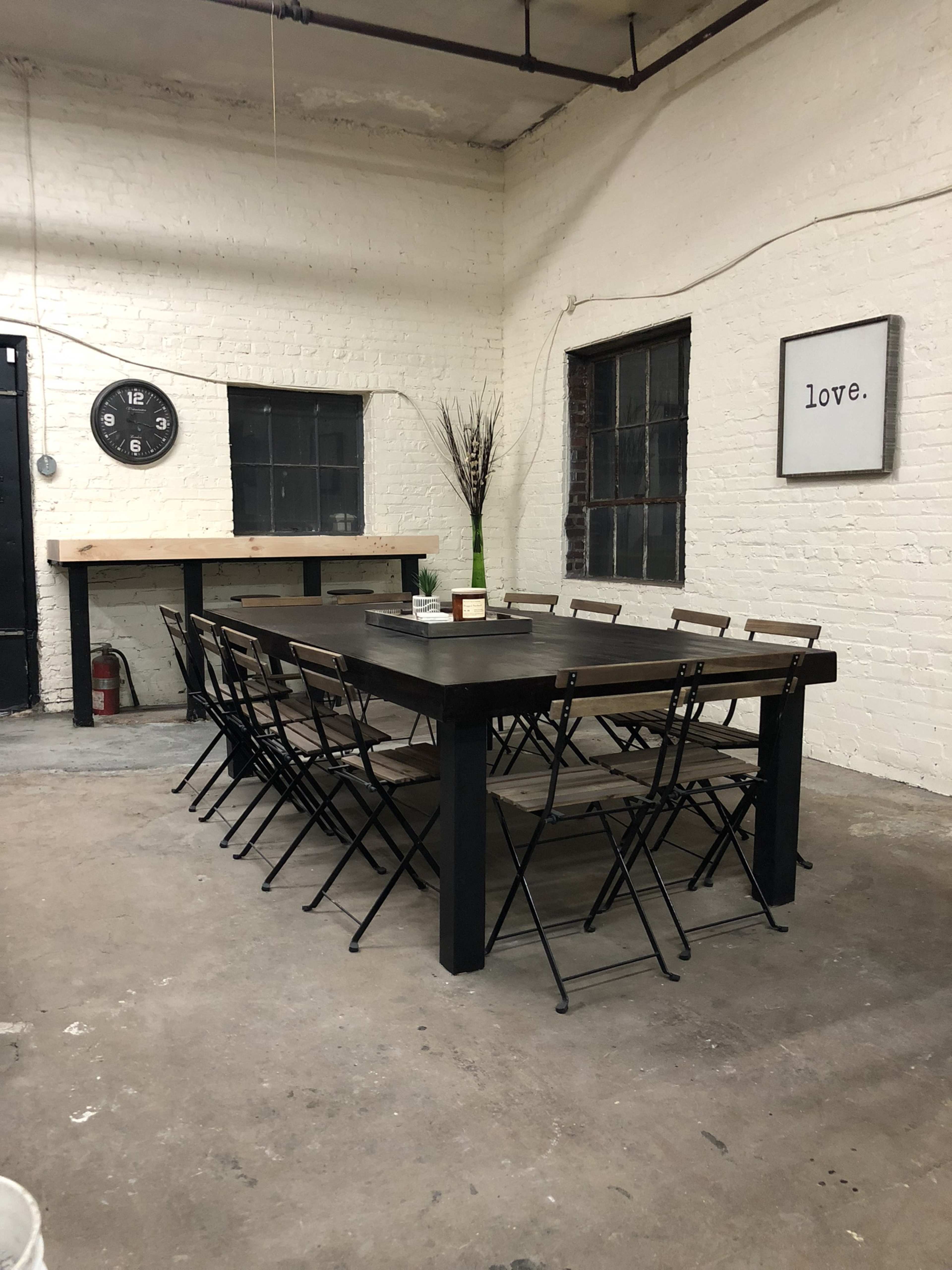 A long wooden table with eight folding chairs is set up in a bare room with white brick walls and large windows.