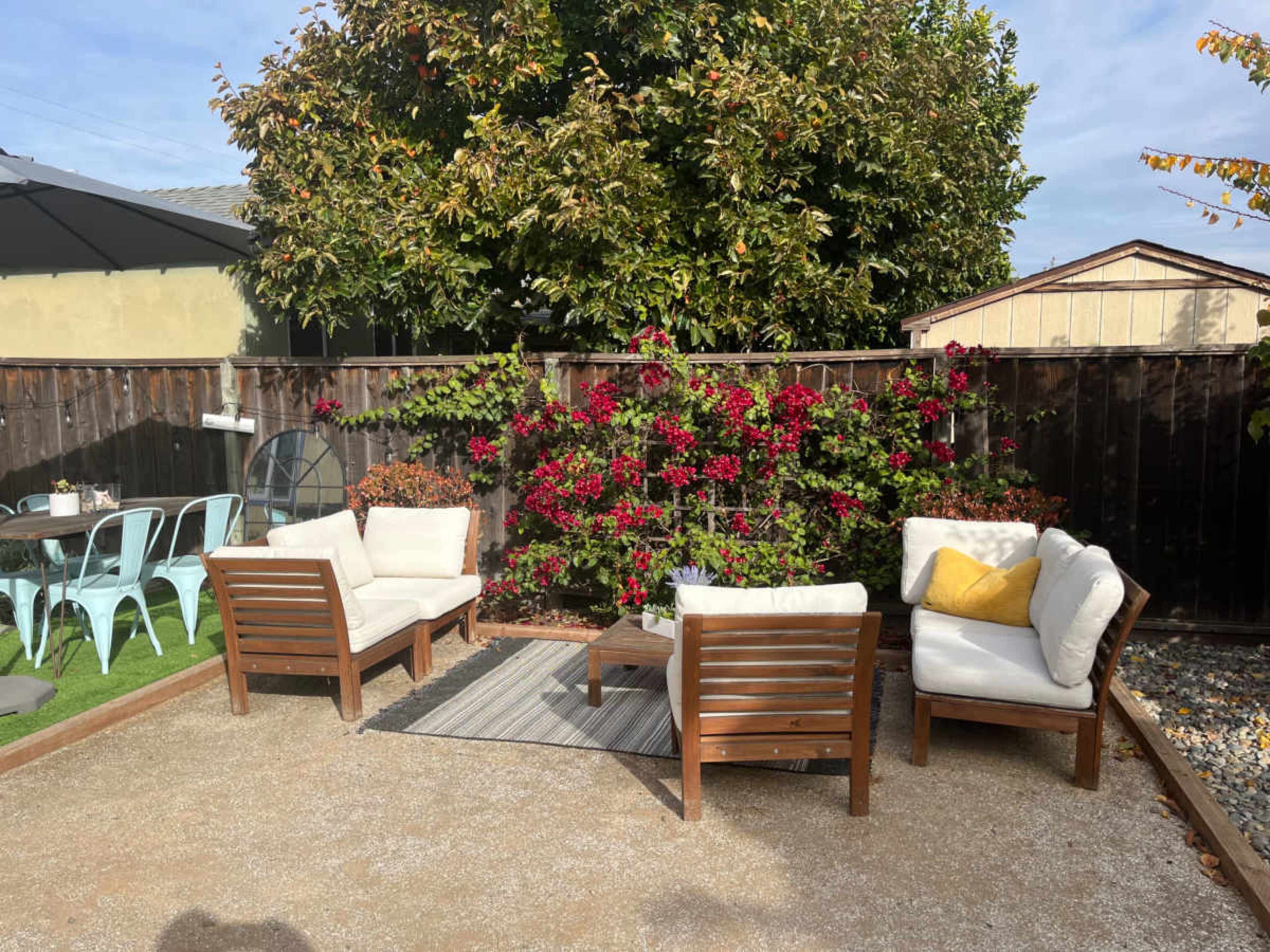 A cozy outdoor seating area features four wooden chairs and a table, surrounded by gravel and vibrant bougainvillea along a fence.
