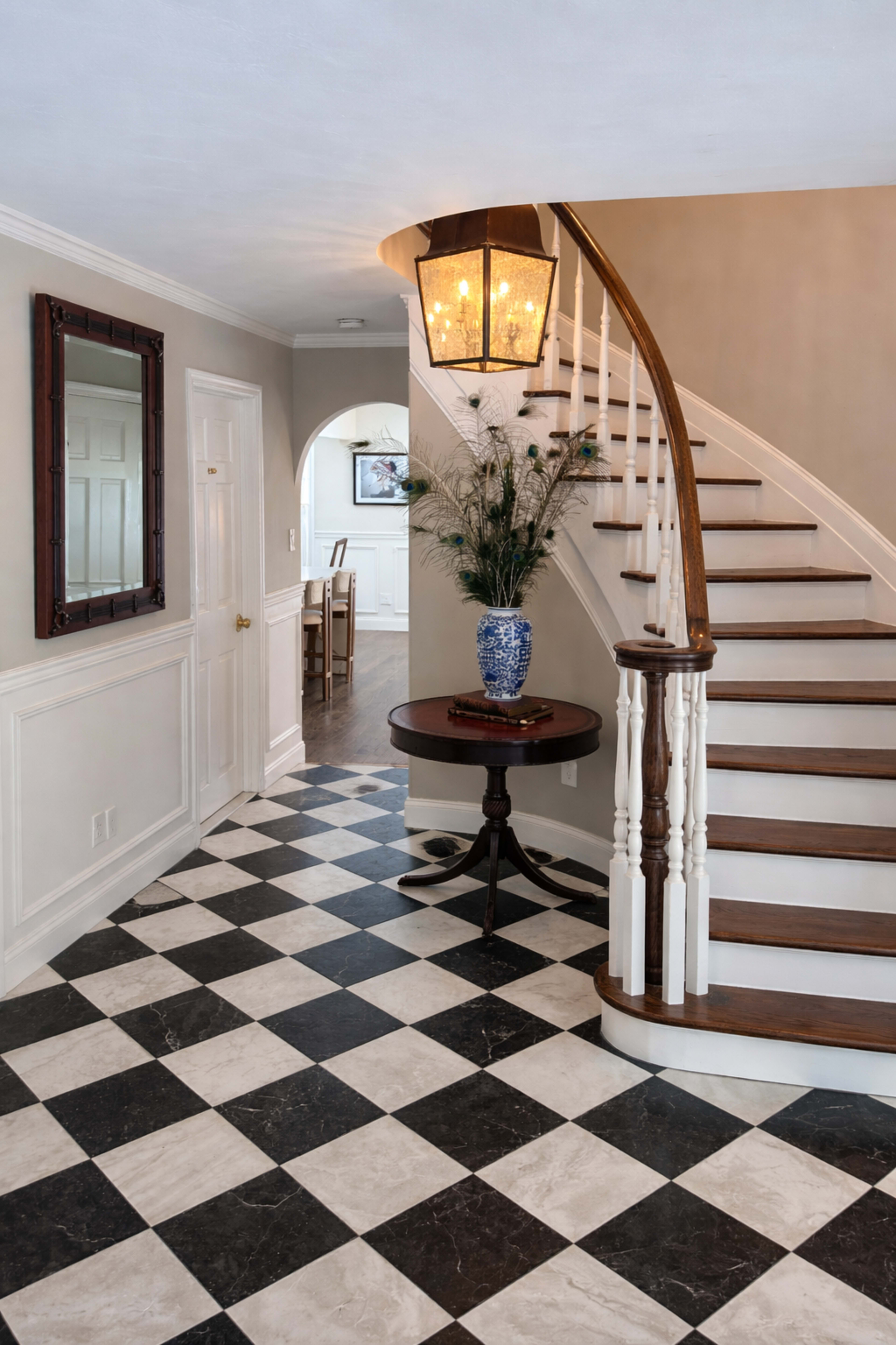 The image shows a bright foyer with a staircase curving to the left, a round table with a blue vase, and a checkered black and white tile floor.