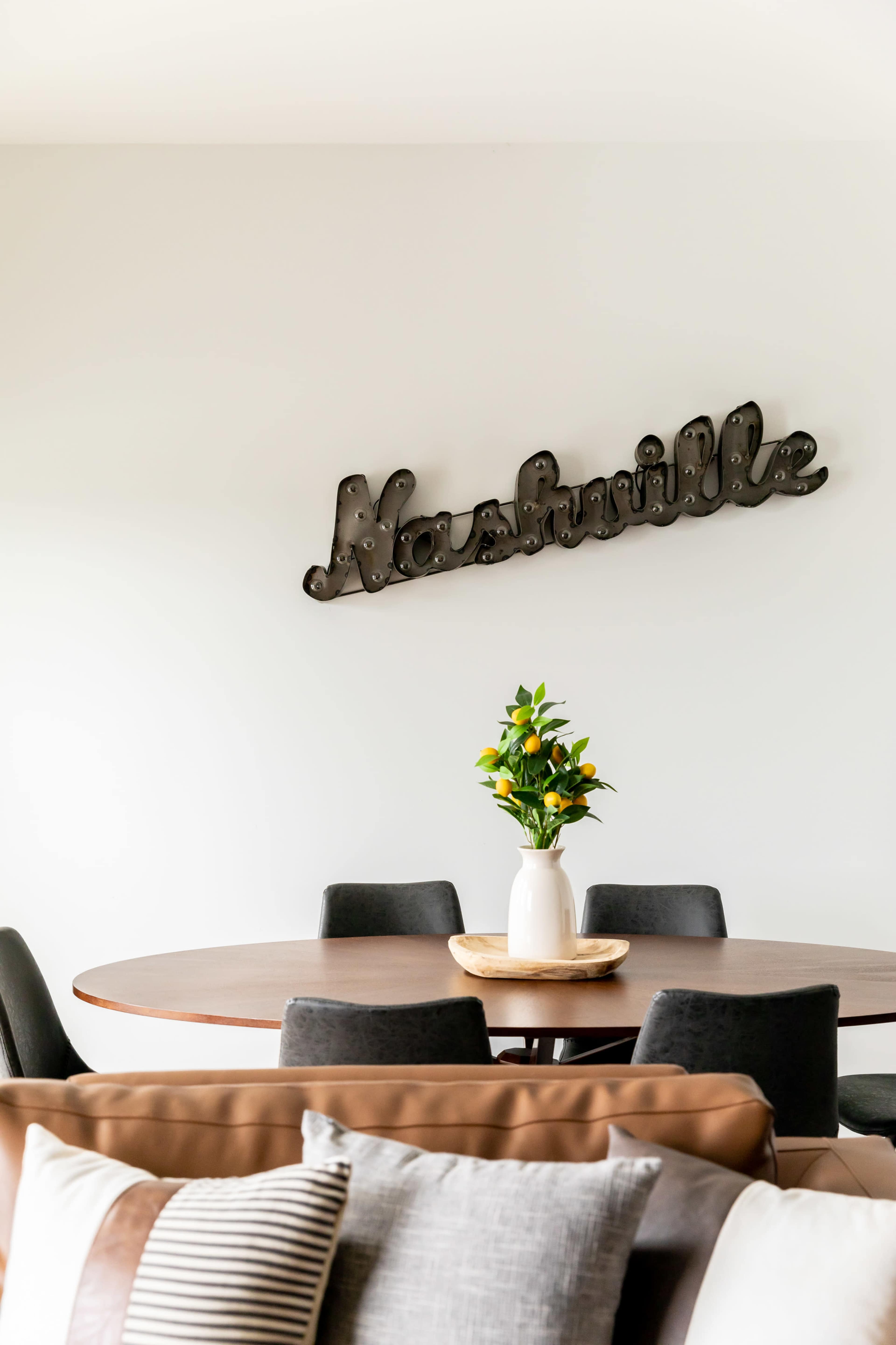A round wooden dining table surrounded by black chairs features a vase of flowers in the center, with a "Nashville" sign mounted on the white wall above.